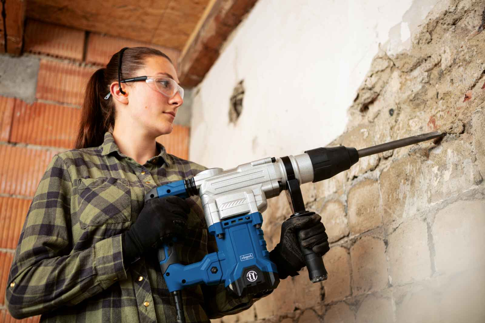 Een vrouw met een veiligheidsbril gebruikt een blauwe sloophamer op een bakstenen muur.