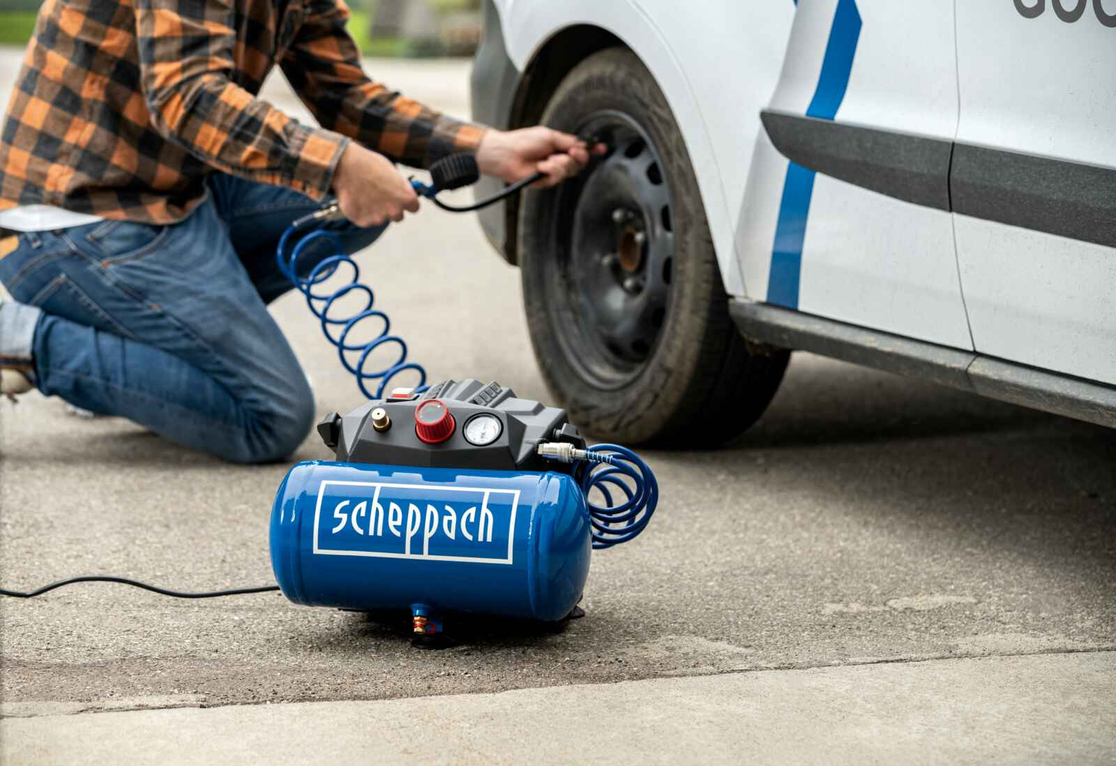 Een man knielt en pompt een band op. Een blauwe luchtcompressor met witte letters staat op de grond.