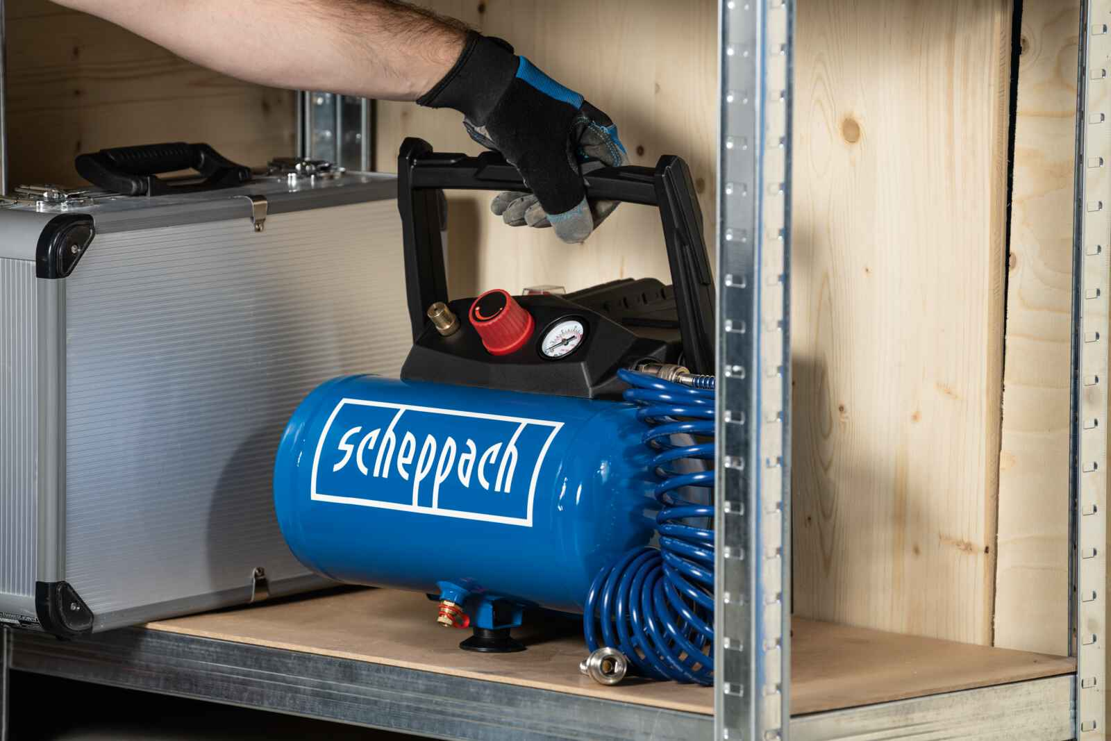 Een hand houdt een blauwe luchtcompressor vast. De compressor heeft een zwarte bovenkant met meters en een wit logo.