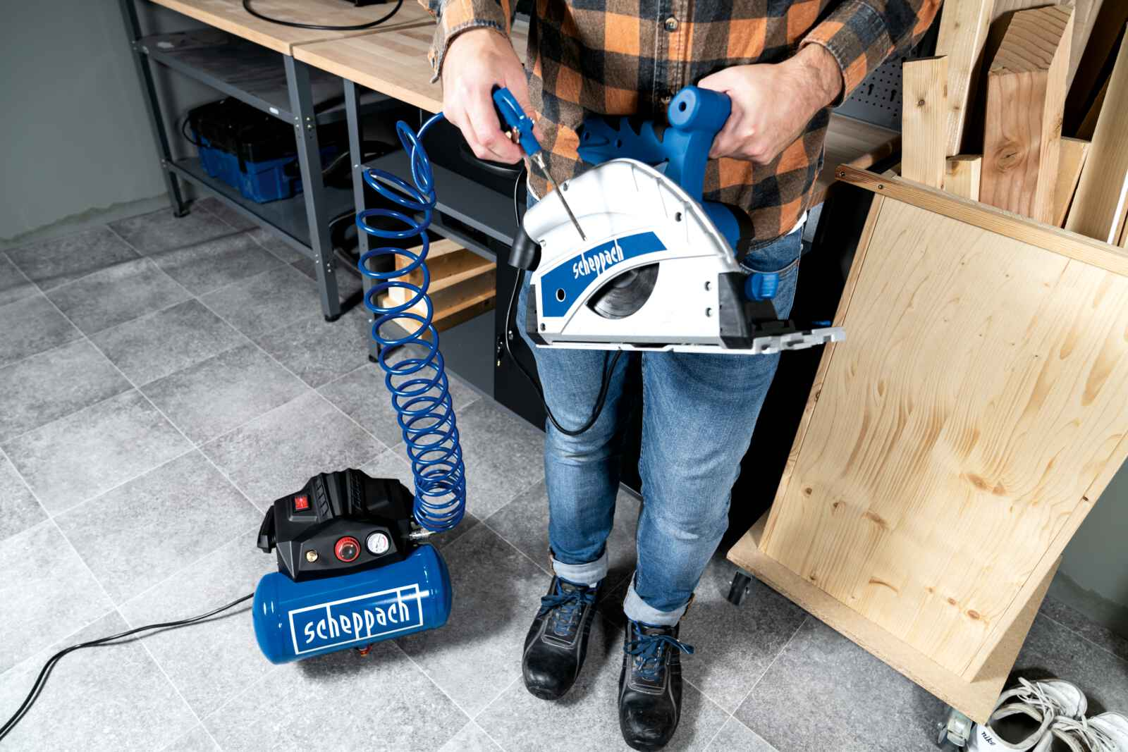 Een man in een geruit shirt en jeans houdt een cirkelzaag en een compressor vast in een werkplaats.