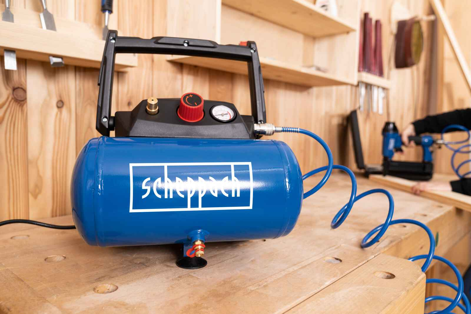 Een blauwe luchtcompressor op een houten tafel. Hij heeft een zwarte bovenkant, een rode wijzerplaat en een wit logo.
