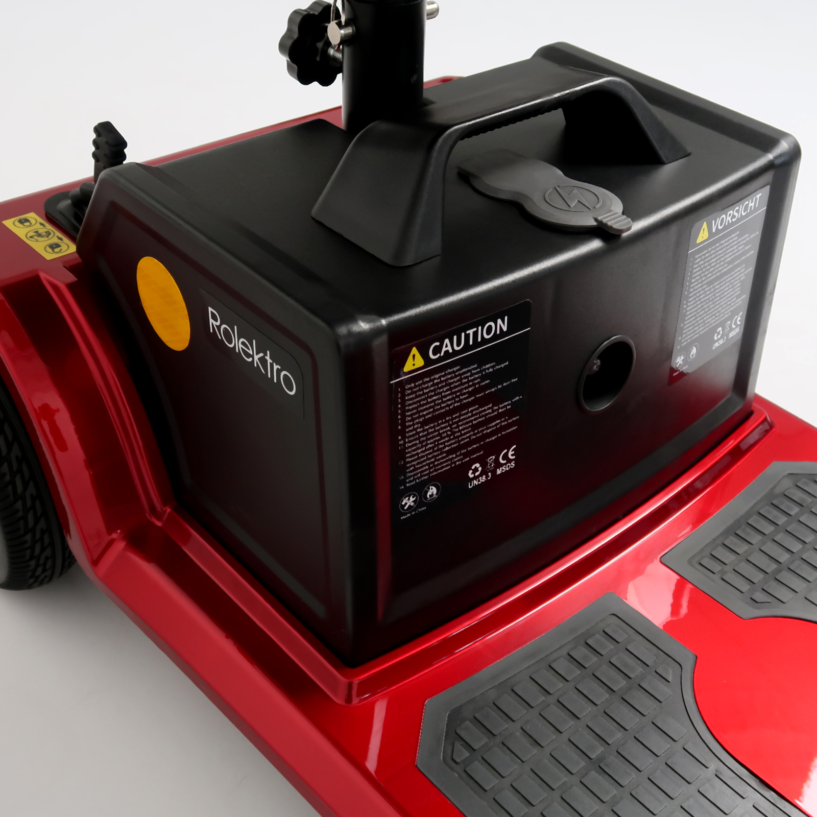 Nahaufnahme eines roten Mobilitätsrollers. Schwarzer Batteriekasten mit Warnhinweisen, gelbes Logo und graue Fußstützen.