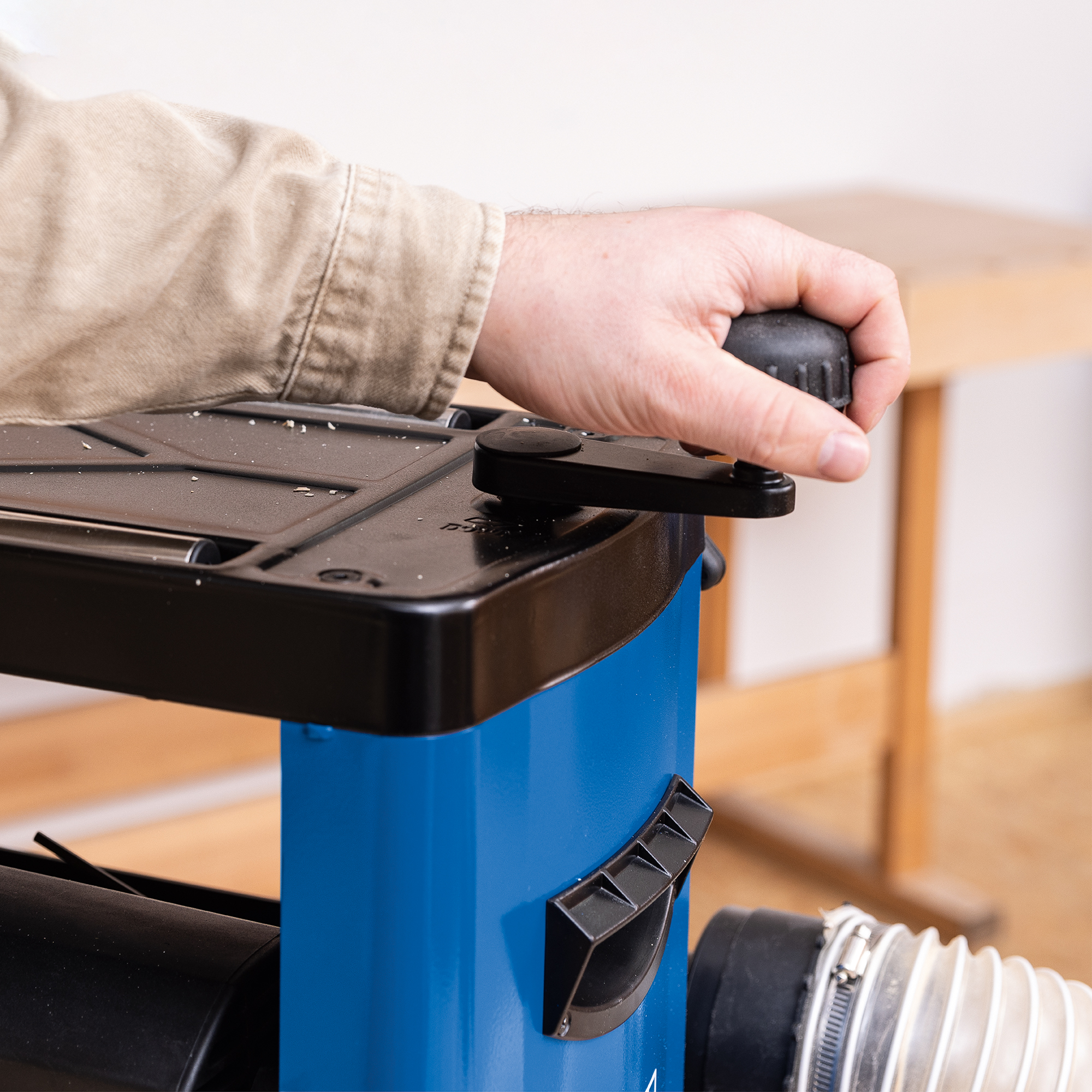 Een hand draait aan een zwarte knop op een blauw-zwarte schaafmachine. De schaafmachine heeft een houten basis.