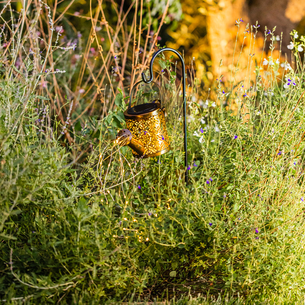 Eine goldene Gießkanne hängt an einem schwarzen Haken inmitten von grünen Pflanzen und Blumen. Szene im Garten.
