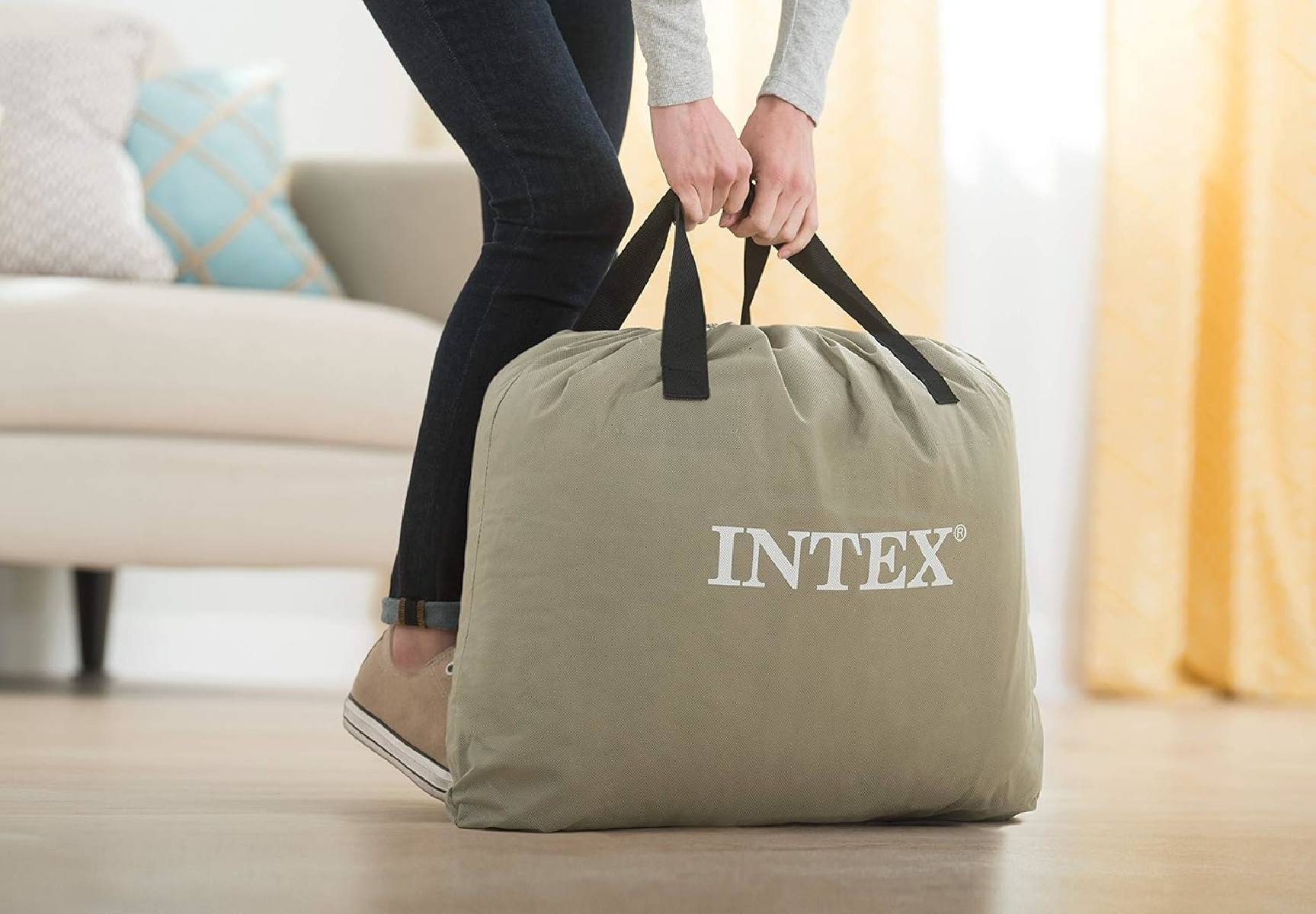 Una persona sostiene una gran bolsa verde con asas negras. La bolsa tiene el logo 'INTEX'. La persona lleva vaqueros y zapatillas.