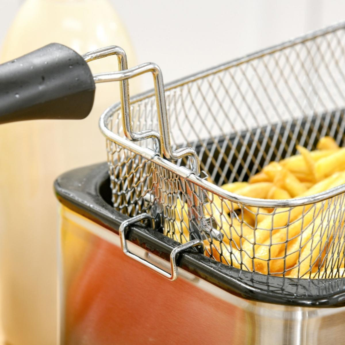 Ein Metallkorb mit Pommes Frites in einer Fritteuse. Eine Flasche Soße ist im Hintergrund. Der Fokus liegt auf den Pommes.