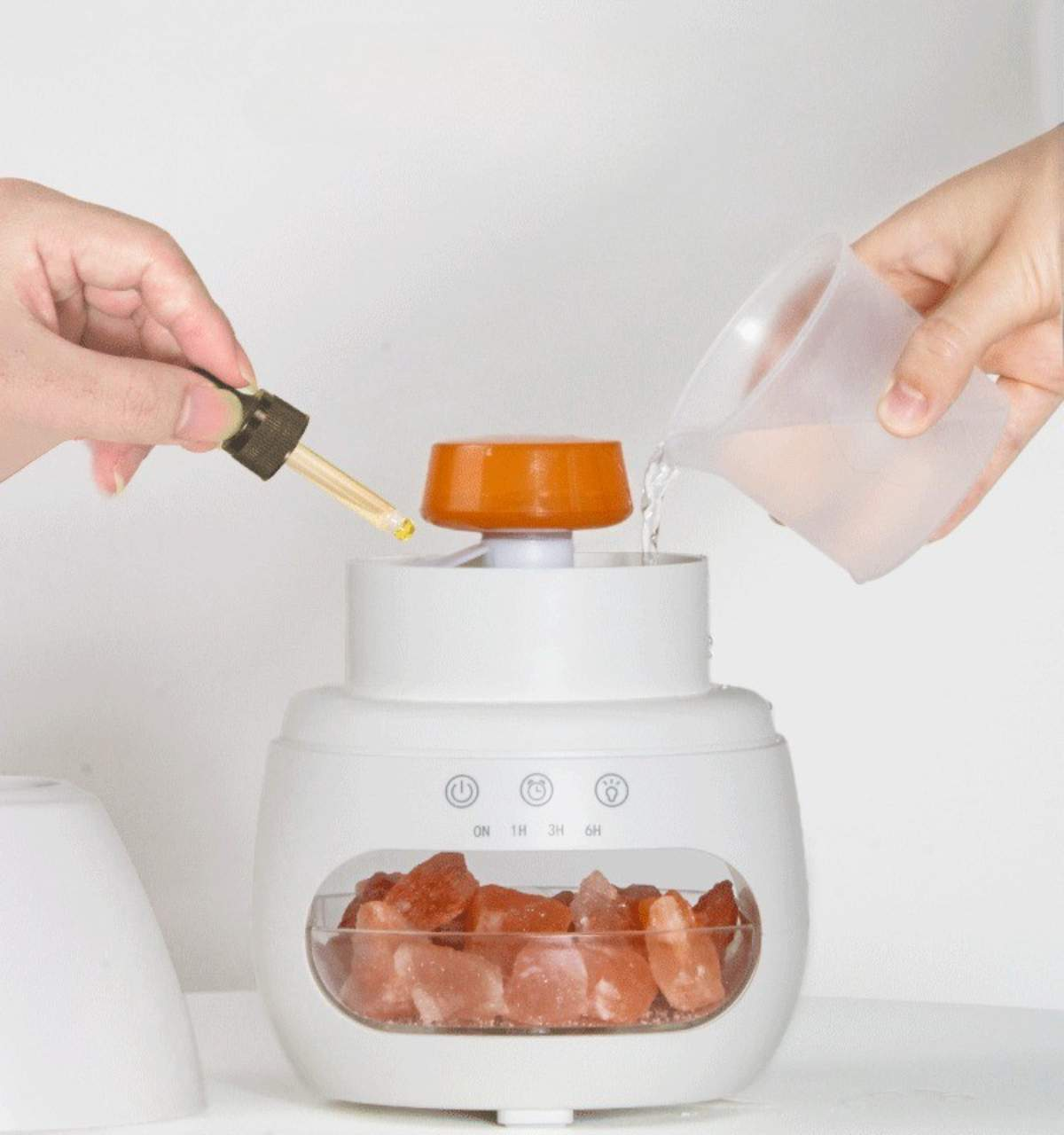 Hände gießen Wasser ein und fügen Tropfen zu einem weißen Luftbefeuchter mit Himalaya-Salz hinzu.