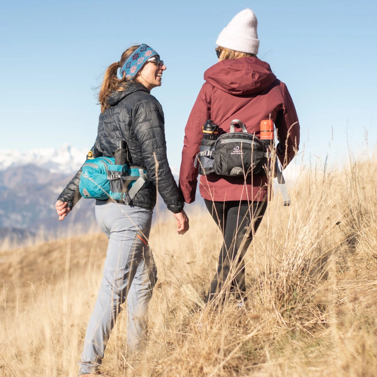 Dos personas caminando con riñoneras. Montañas y árboles en el fondo.