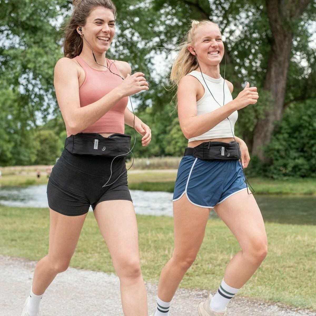 Dos mujeres corriendo con cinturones de running y auriculares. Árboles verdes al fondo.