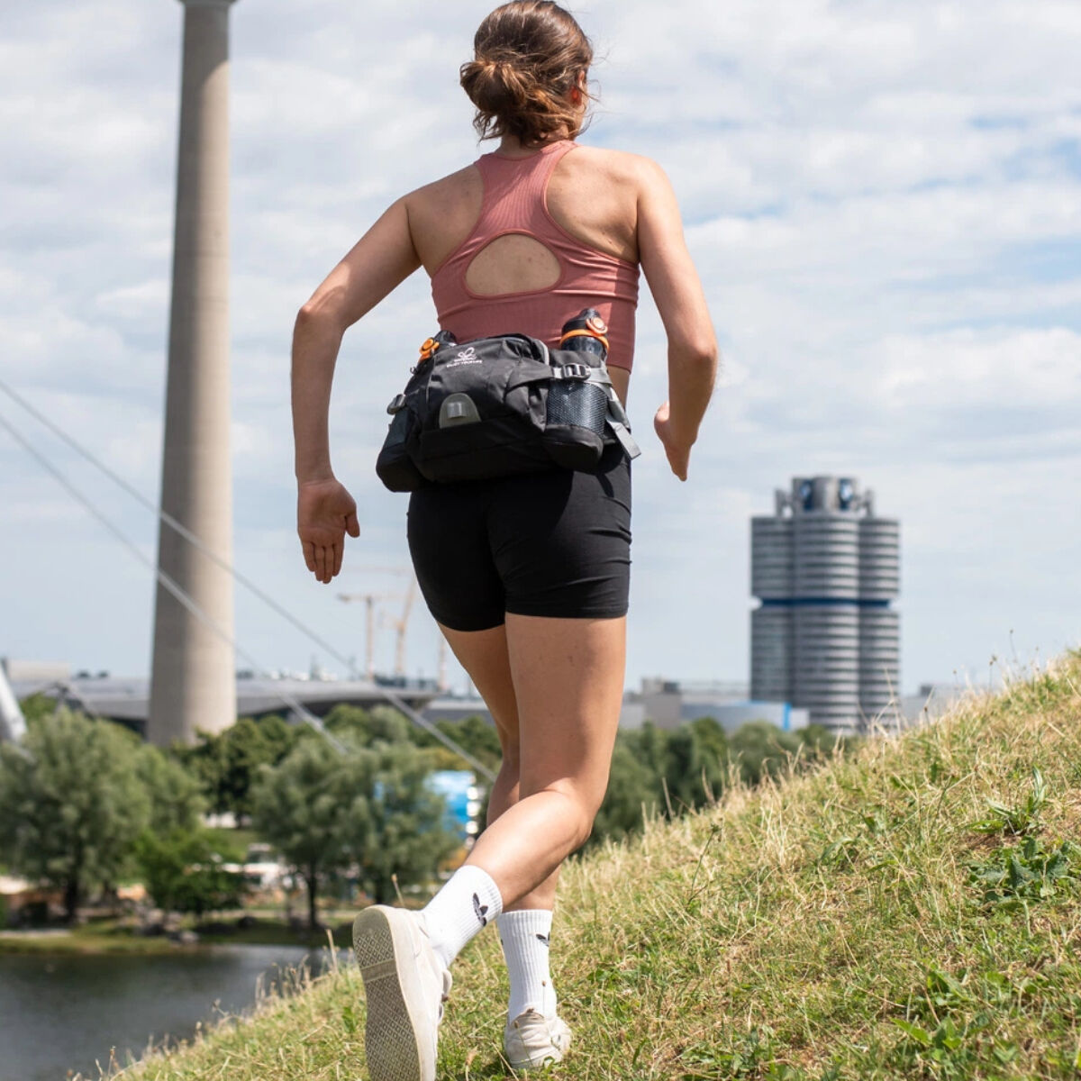 Una mujer corriendo con un cinturón de running. Ciudad y hierba en el fondo.
