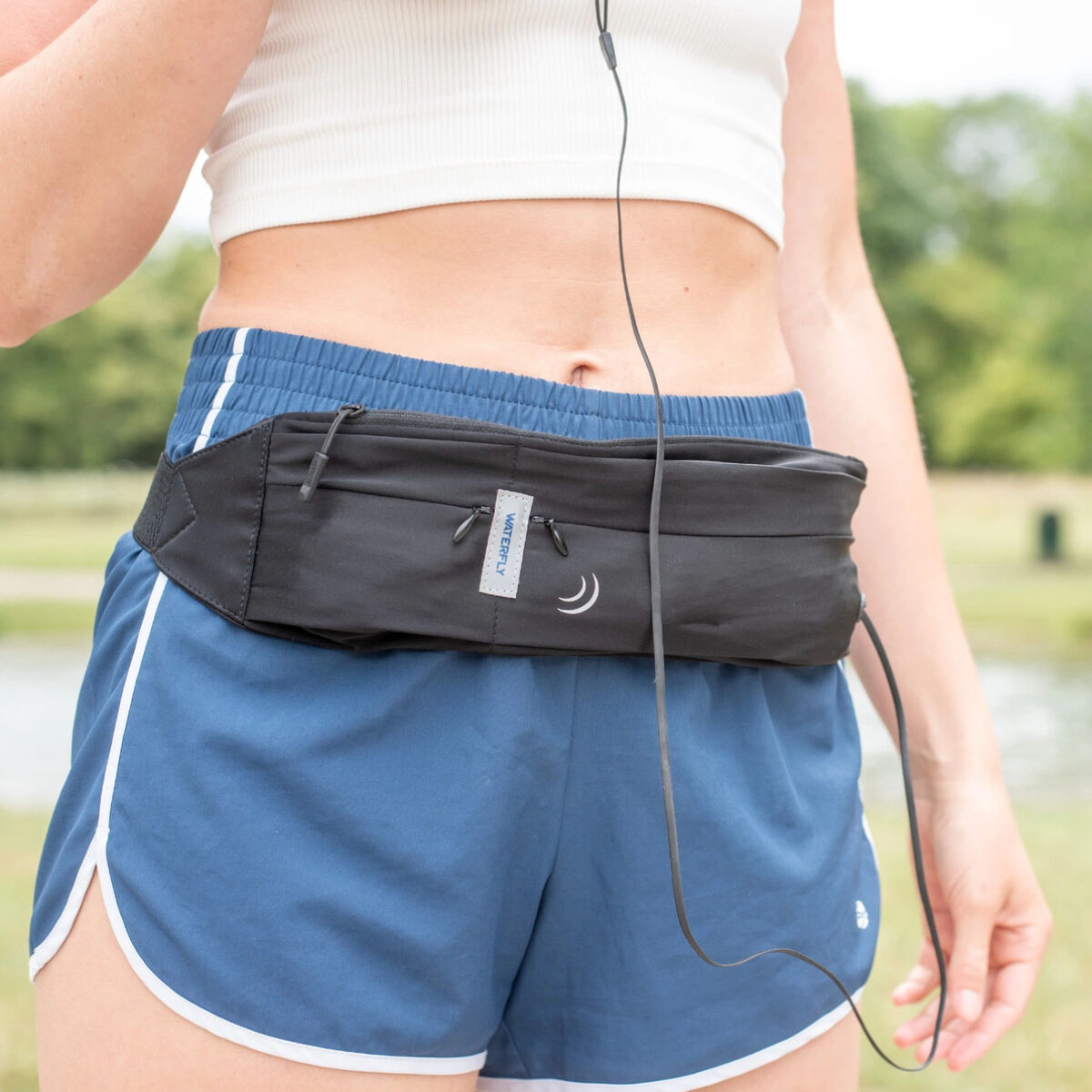 Una persona indossa un top bianco e pantaloncini blu con marsupio nero. Lo sfondo mostra alberi verdi ed erba.