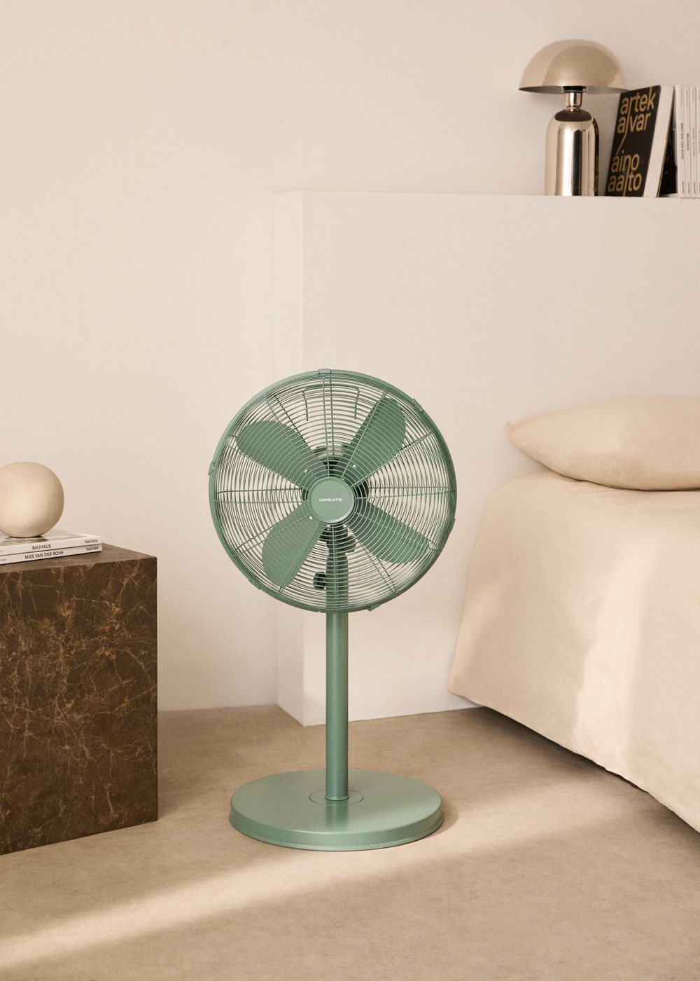 Ventilador de pie verde en una habitación. Está cerca de una cama y una mesa con lámpara.