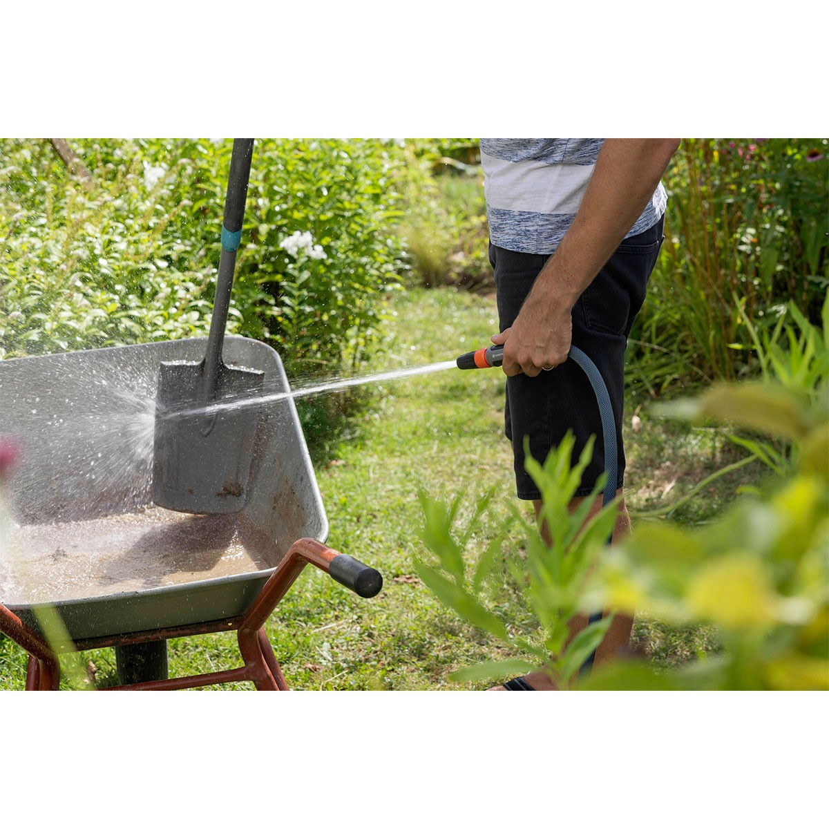 Persona che innaffia una carriola con un tubo. Erba verde e piante sullo sfondo. Acqua limpida.