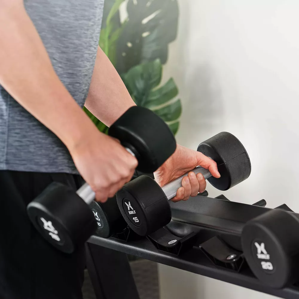 Persoon die twee dumbbells vasthoudt. Zwarte dumbbells. Gym met gewichtenrek. Grijs shirt.