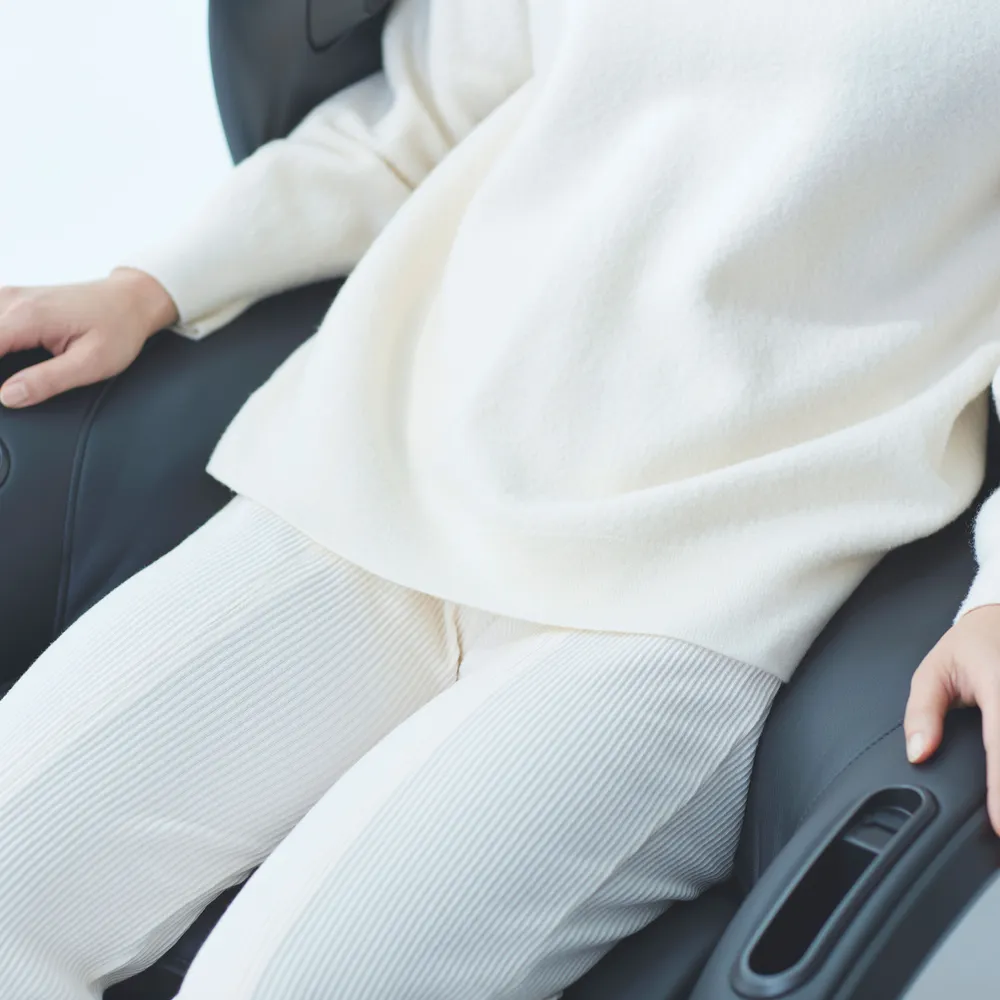 Une personne portant un pull et un pantalon blancs assise dans un fauteuil de massage noir.