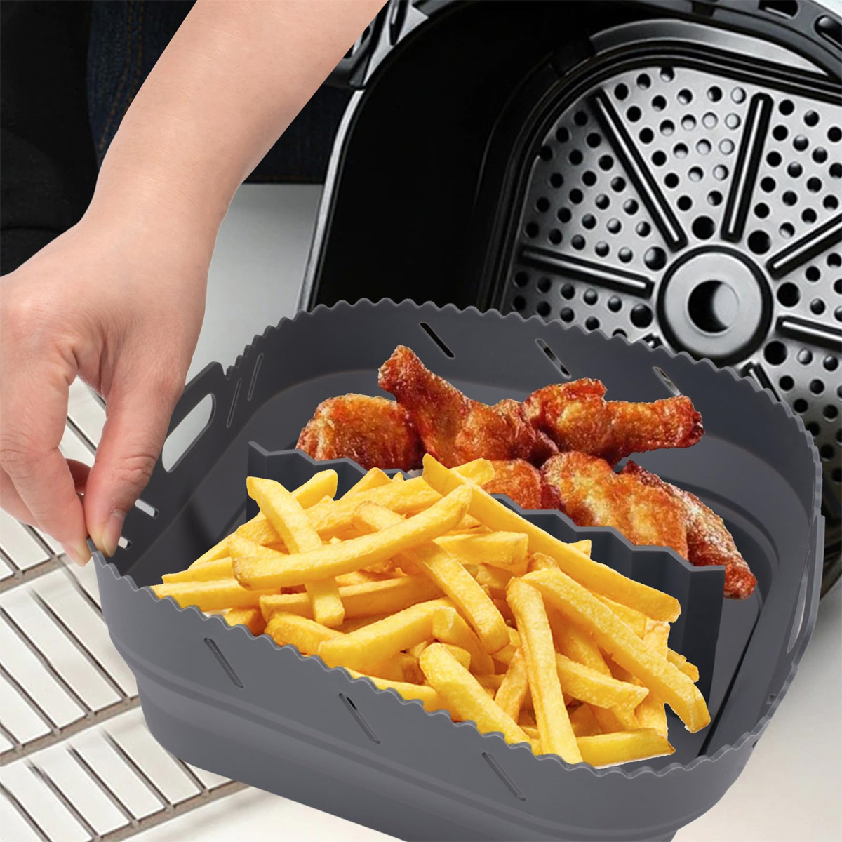 Eine Hand, die ein graues Heißluftfritteusen-Zubehör hält, das mit Pommes Frites und Hähnchenflügeln gefüllt ist. Eine Heißluftfritteuse ist im Hintergrund.
