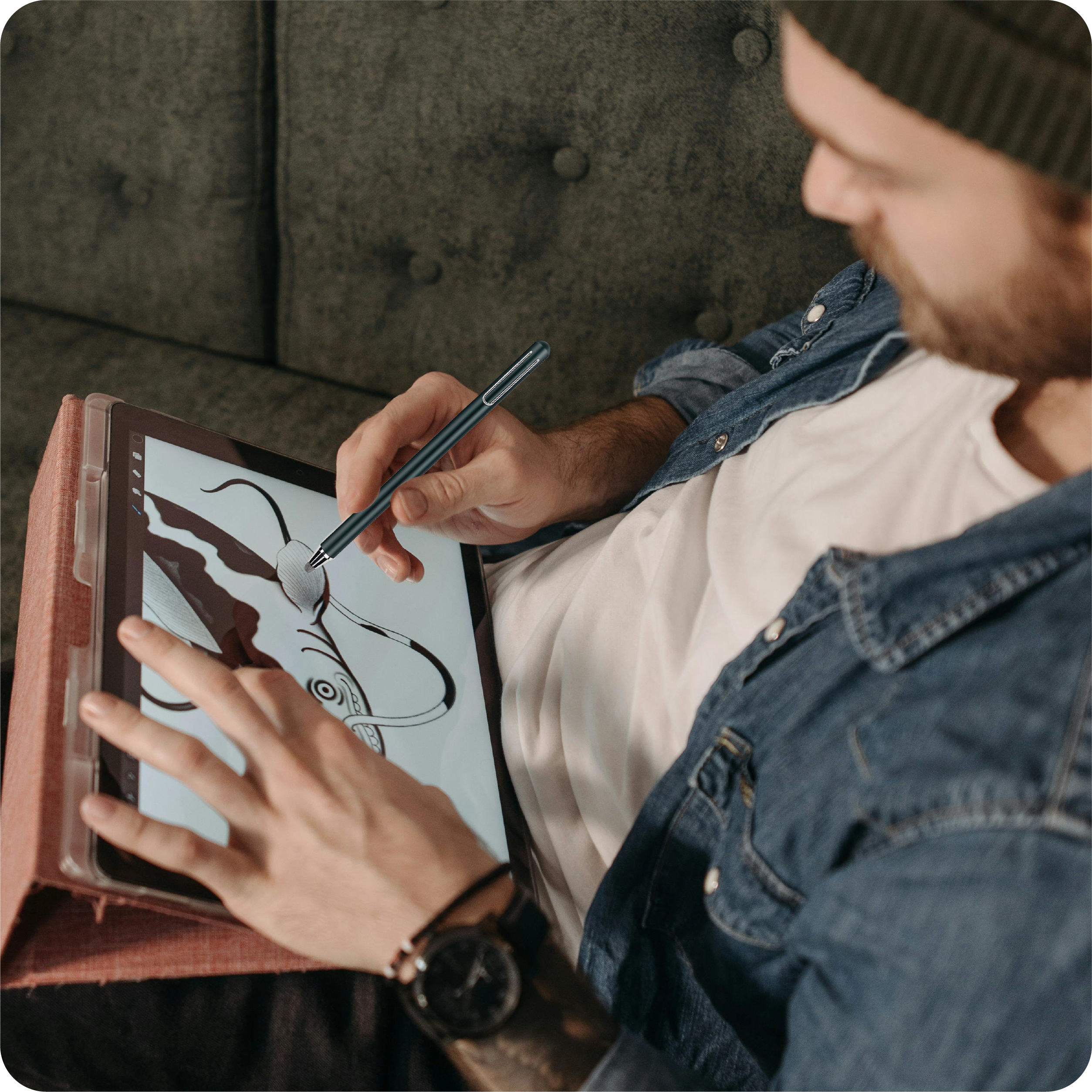 Ein Mann zeichnet mit einem Stift auf einem Tablet. Er sitzt auf einer Couch und trägt eine Jeansjacke und eine Mütze.