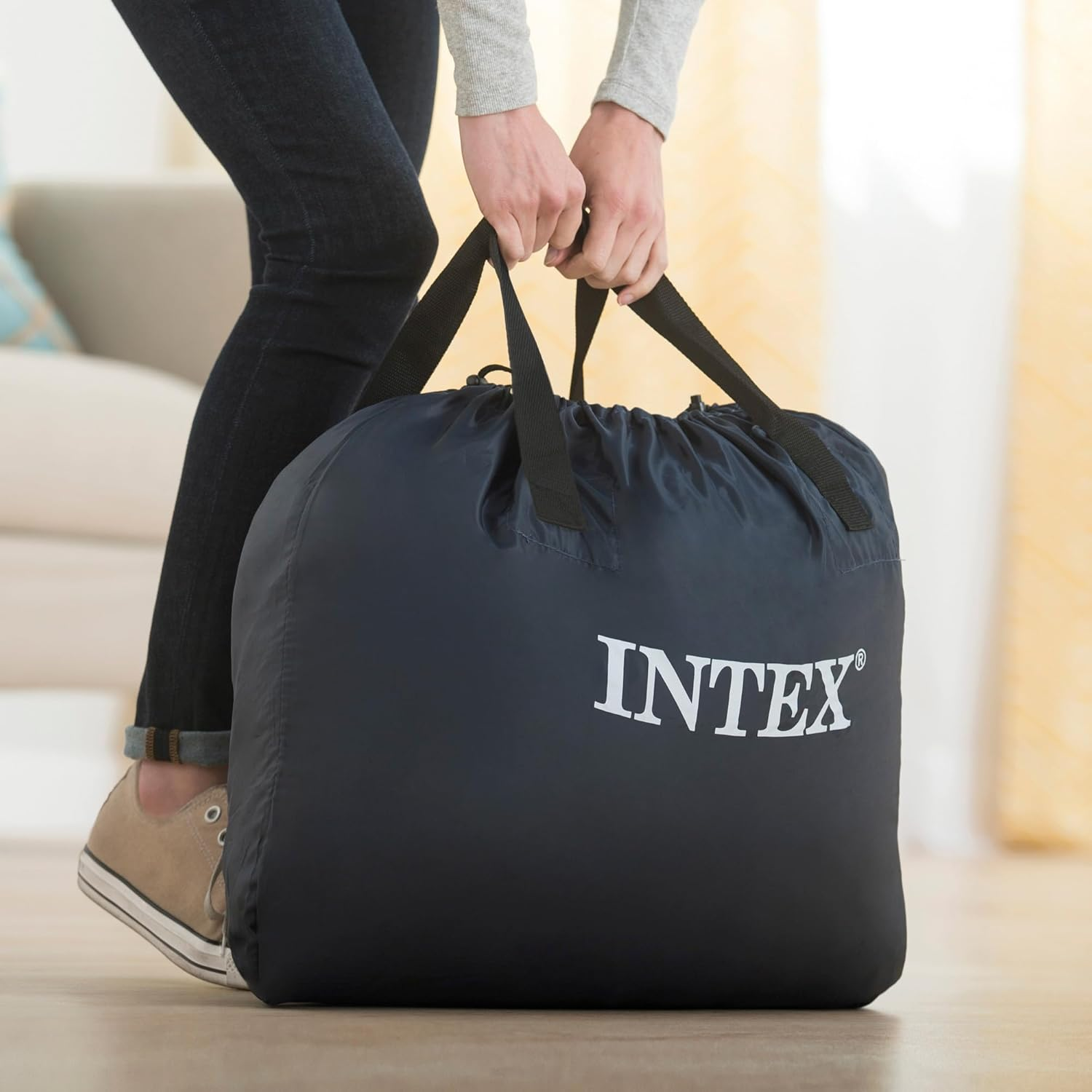 Person hält eine schwarze Intex-Tasche mit weißem Logo, drinnen, auf Holzboden.