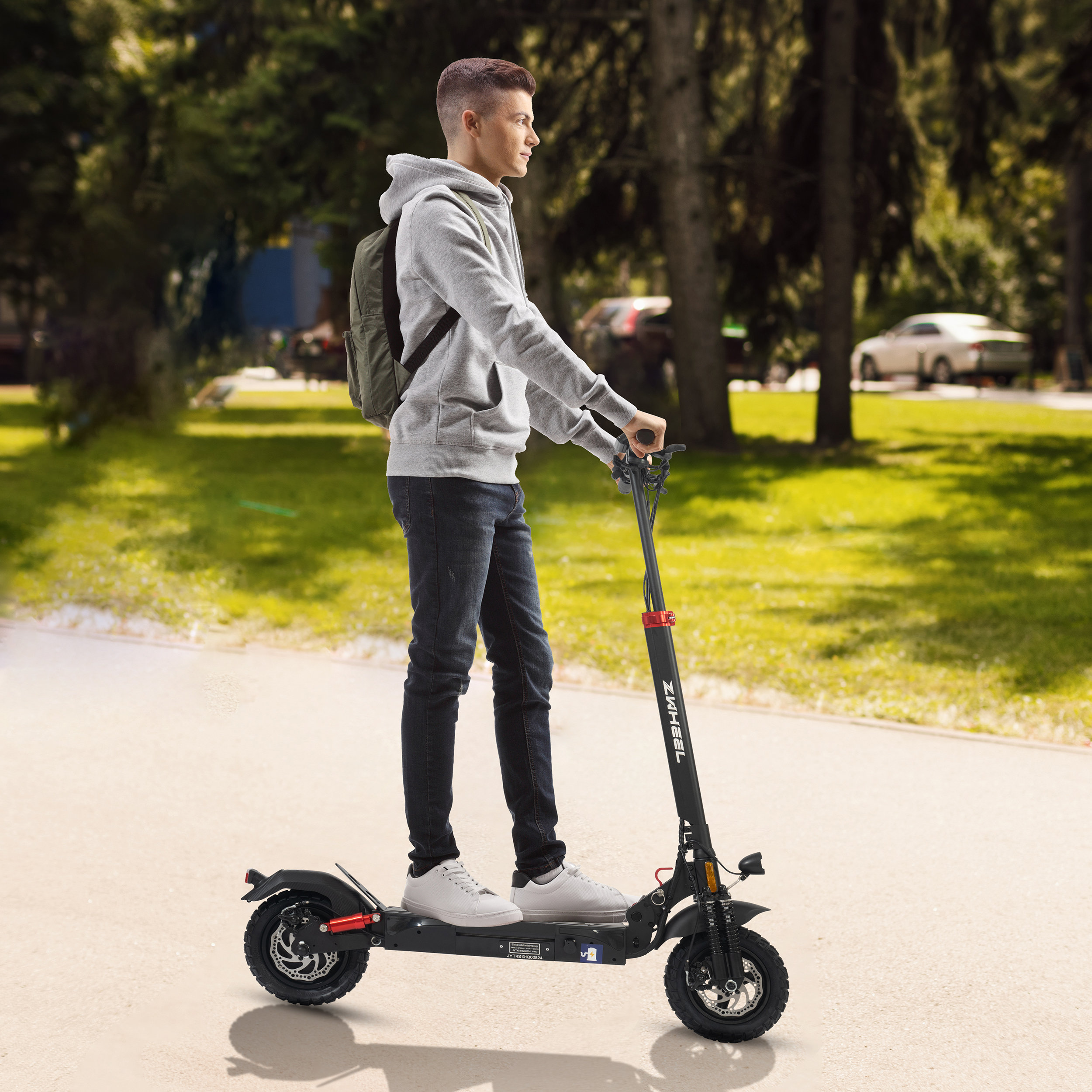 Młody mężczyzna na hulajnodze elektrycznej w parku. Ma na sobie szarą bluzę z kapturem, dżinsy i plecak. Hulajnoga jest czarna.