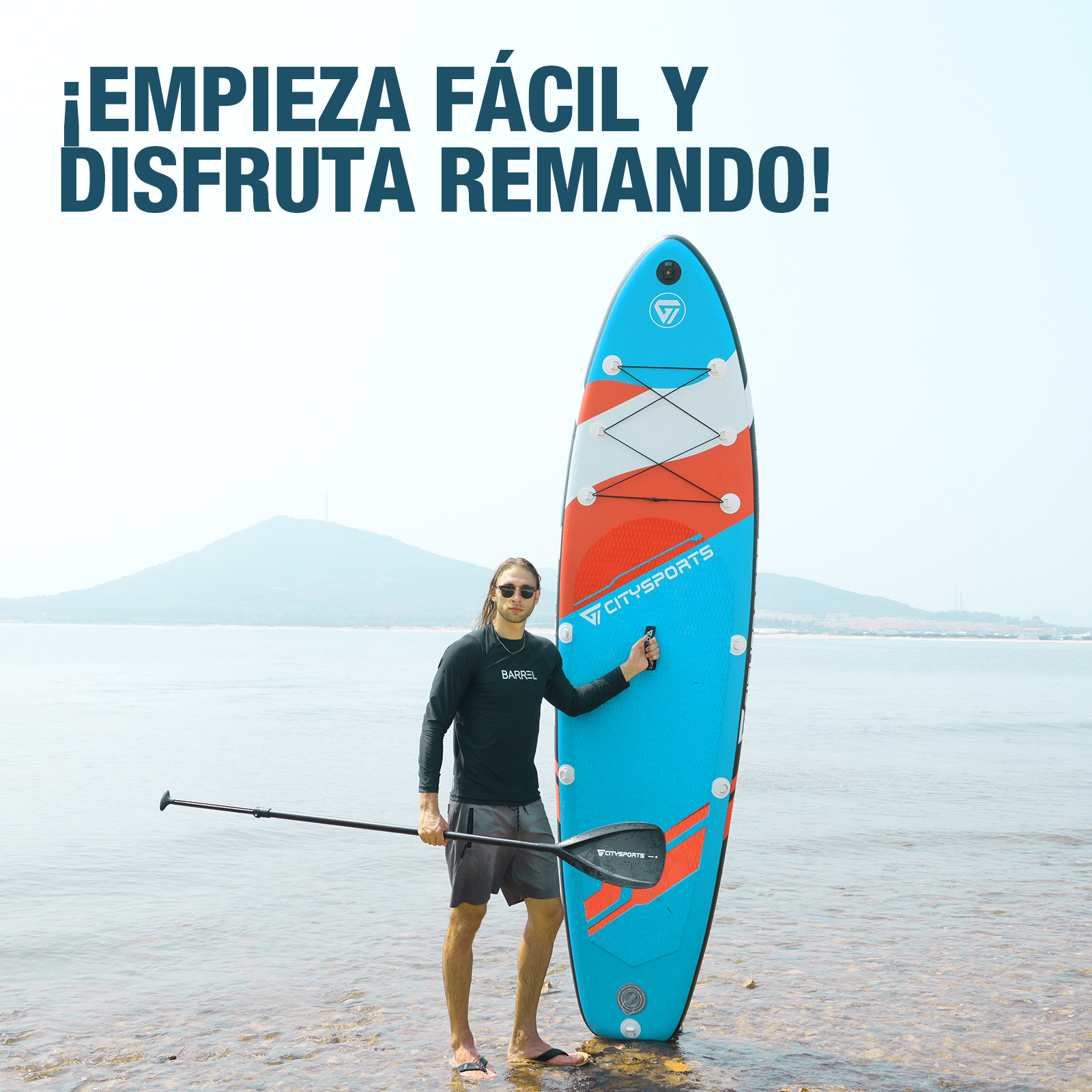 Un hombre está de pie con una tabla de paddle surf en el agua, con una cordillera al fondo.