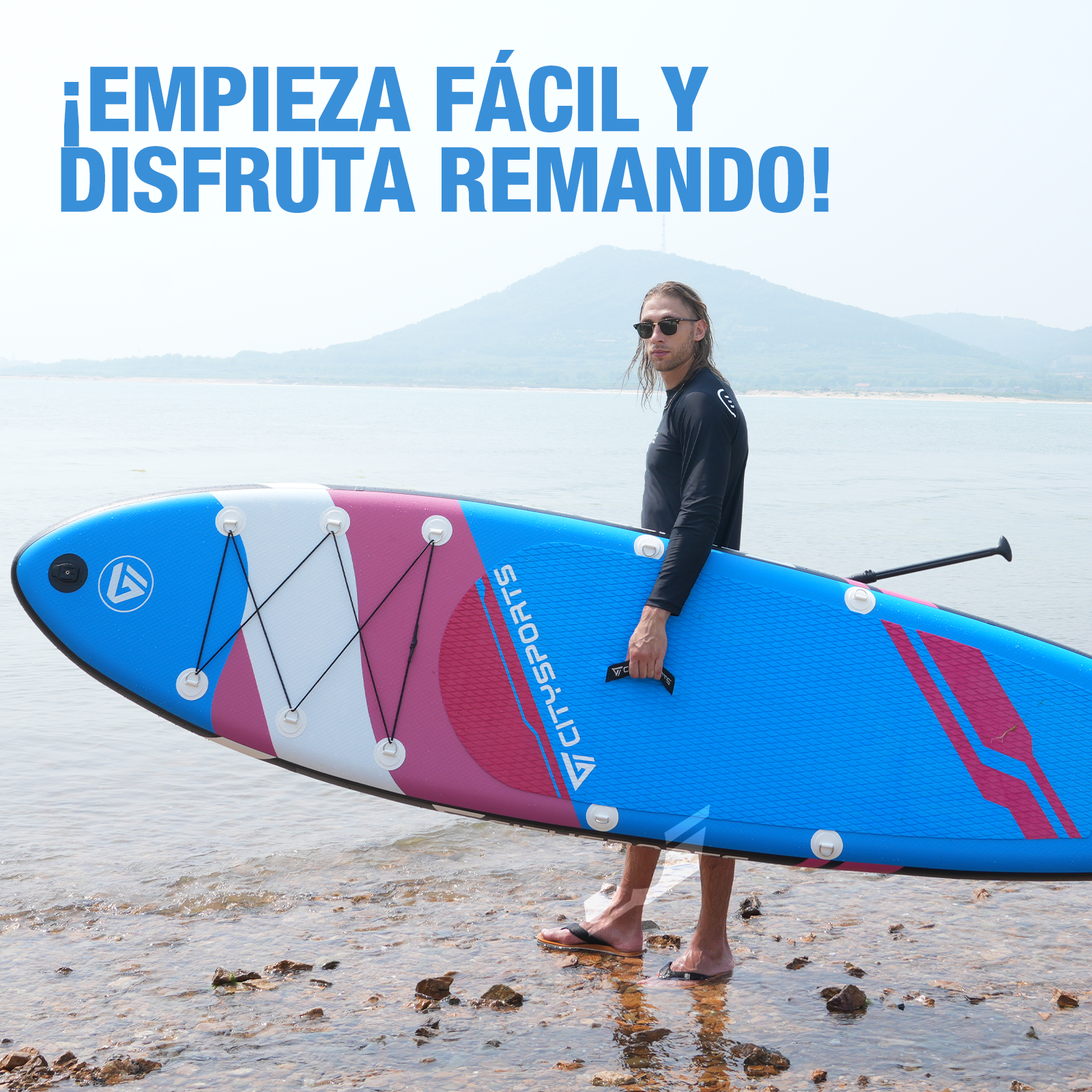 Un hombre sosteniendo una tabla de paddle azul y rosa en una playa. Una montaña y agua son visibles en el fondo.