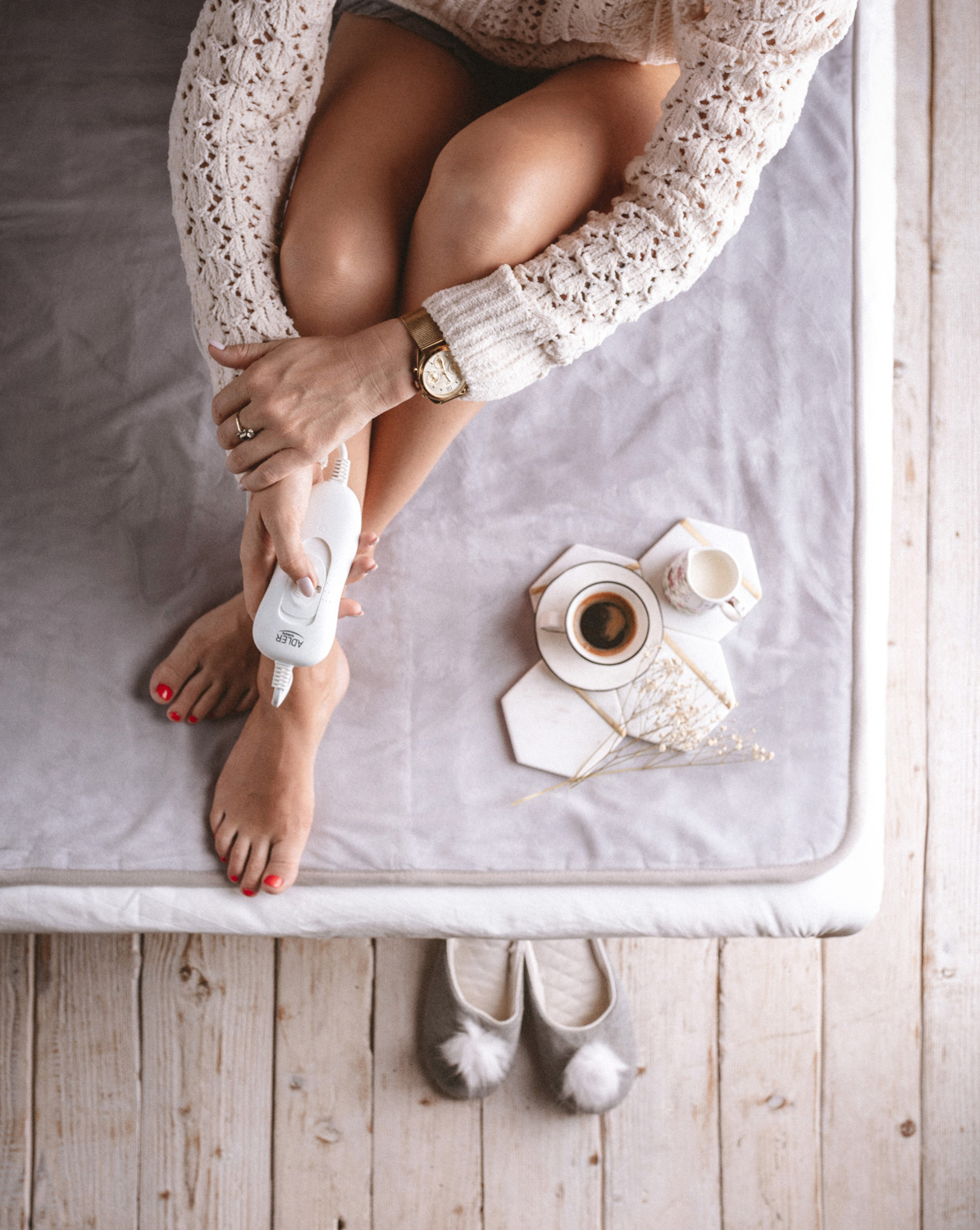 Kobieta na łóżku, z kawą i kapciami. Trzyma urządzenie do ogrzewania stóp.