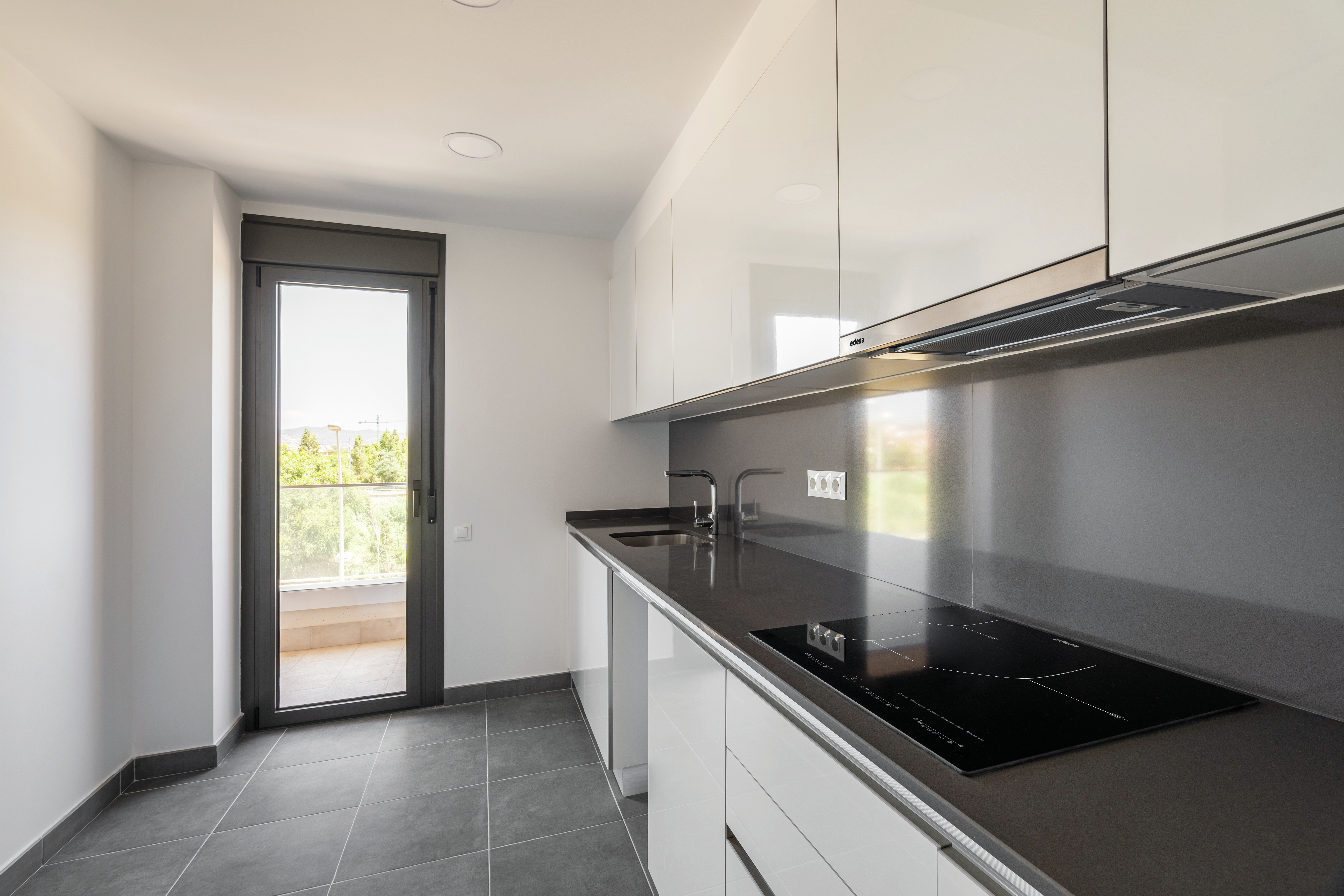 Cocina moderna con gabinetes blancos, encimera negra y puerta de vidrio a balcón con vegetación.