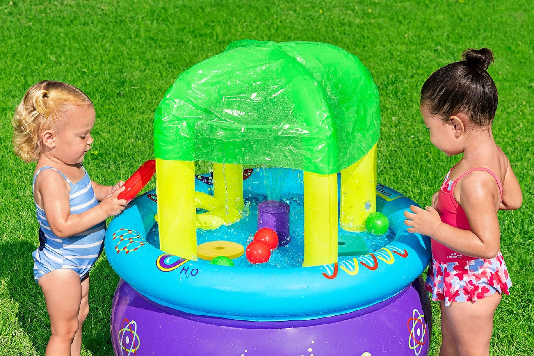 Due bambini piccoli giocano con un tavolo d'acqua sull'erba. Il tavolo ha un tetto verde e spruzza acqua. All'interno ci sono giocattoli e palline.