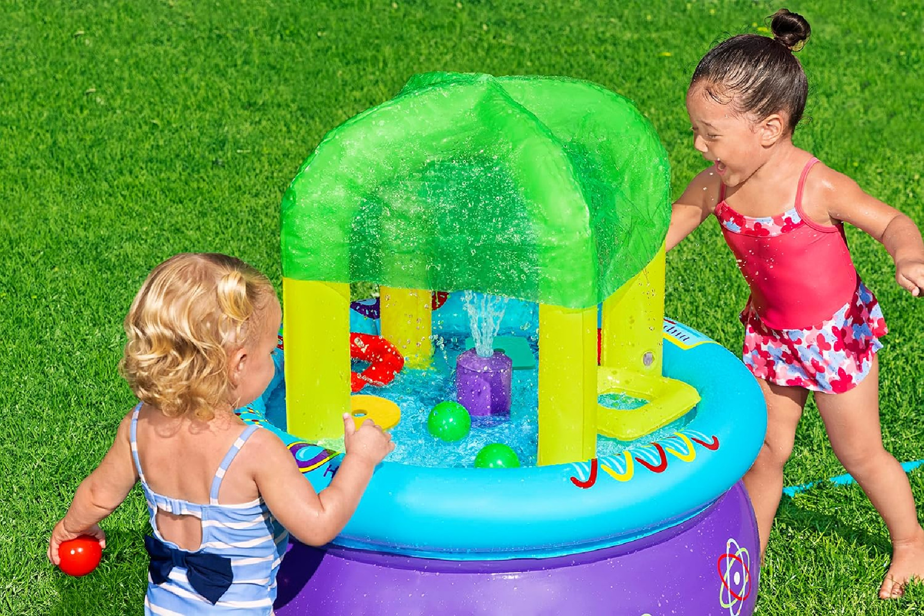 Due ragazze giocano in un tavolo d'acqua sull'erba. Il tavolo ha una tettoia verde e una fontana. Una indossa il rosso, l'altra il blu.