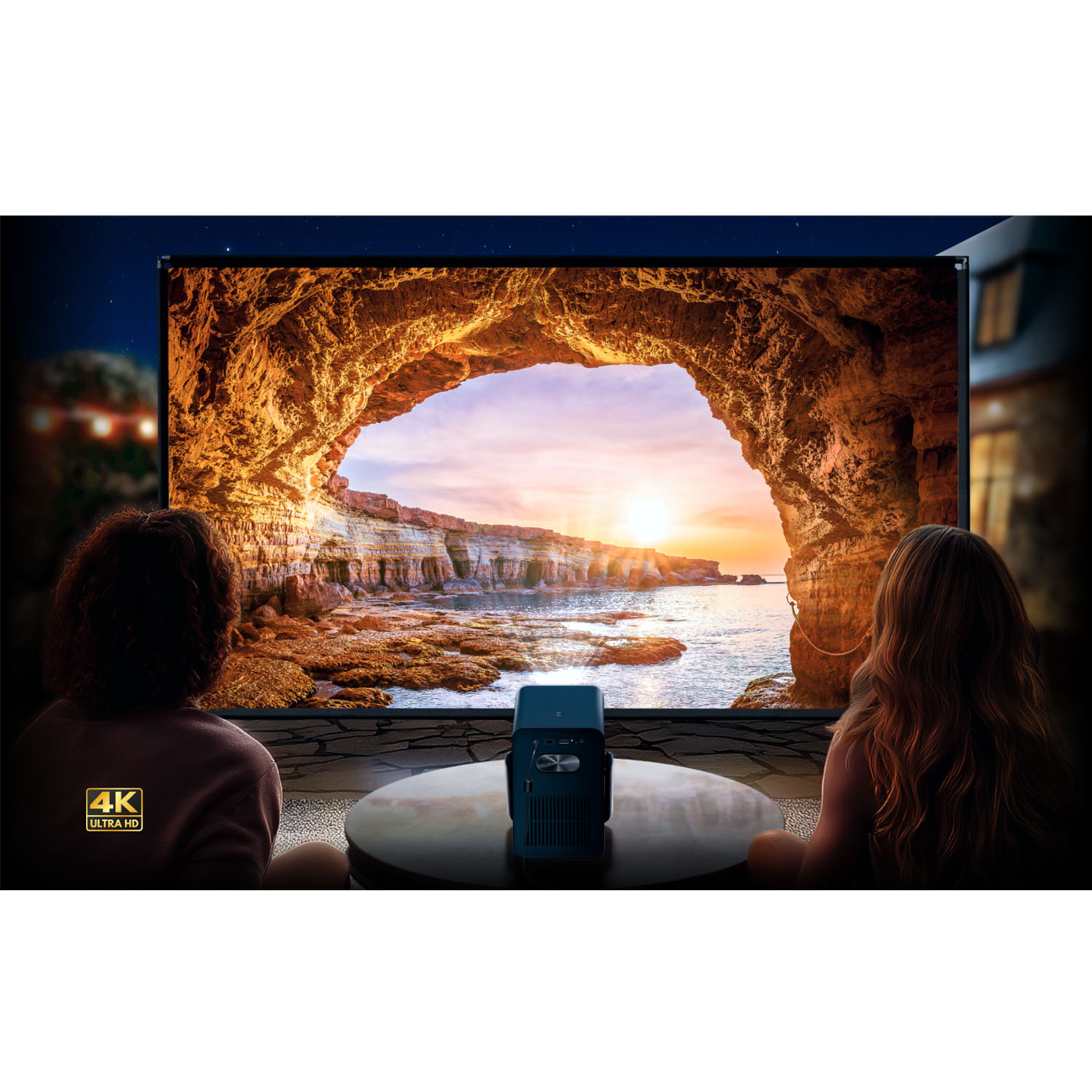 Dwie osoby oglądają wyświetlany obraz zachodu słońca nad brzegiem morza. Projektor stoi na stole. Logo 4K Ultra HD.