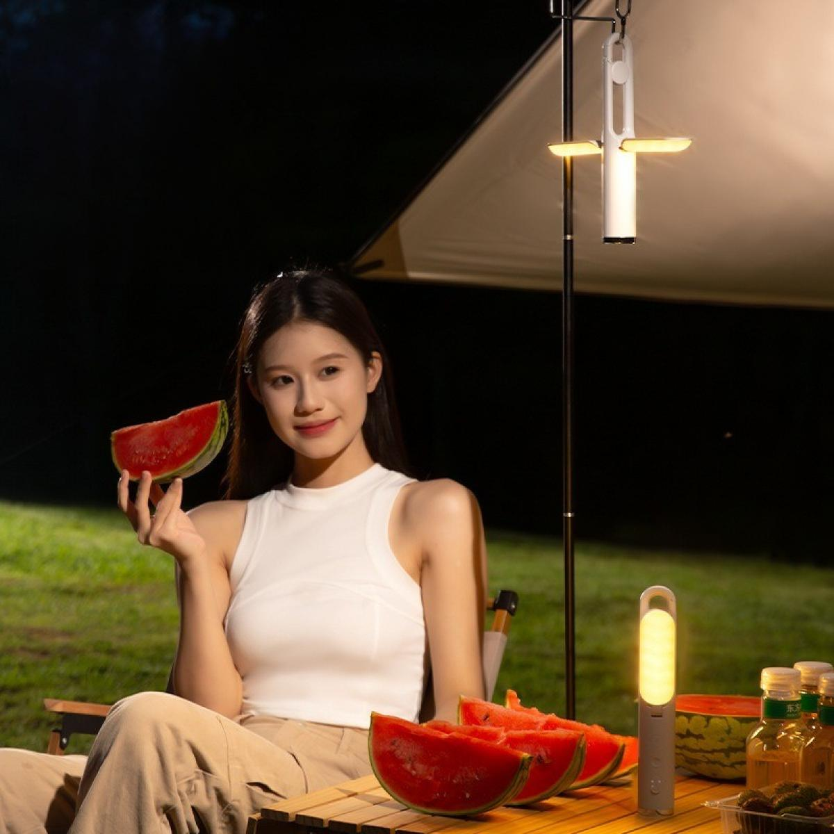Eine Frau hält Wassermelone. Scheiben auf einem Tisch. Camping-Szene mit Lampe und Sonnenschirm.