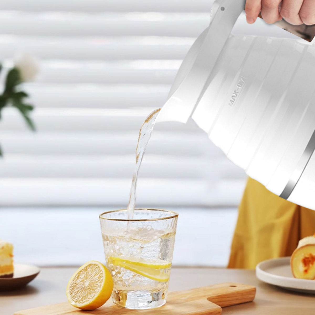 Ein weißer Wasserkocher gießt Wasser in ein Glas mit Zitronenscheiben. Eine halbe Zitrone liegt auf einem Holzbrett mit Kuchen in der Nähe.