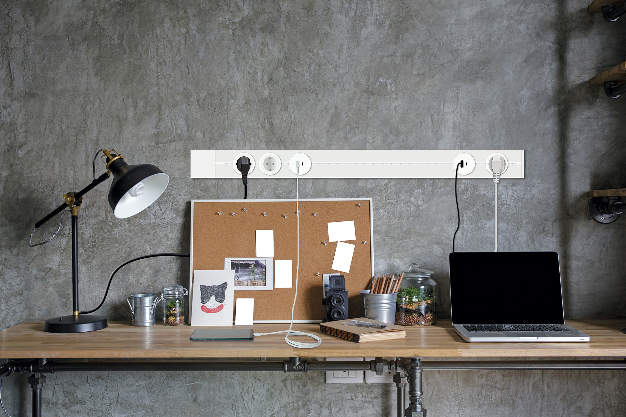 Ein Schreibtisch mit Laptop, Lampe, Pinnwand und verschiedenen Gegenständen. Steckdosenleiste darüber. Betonwand im Hintergrund.