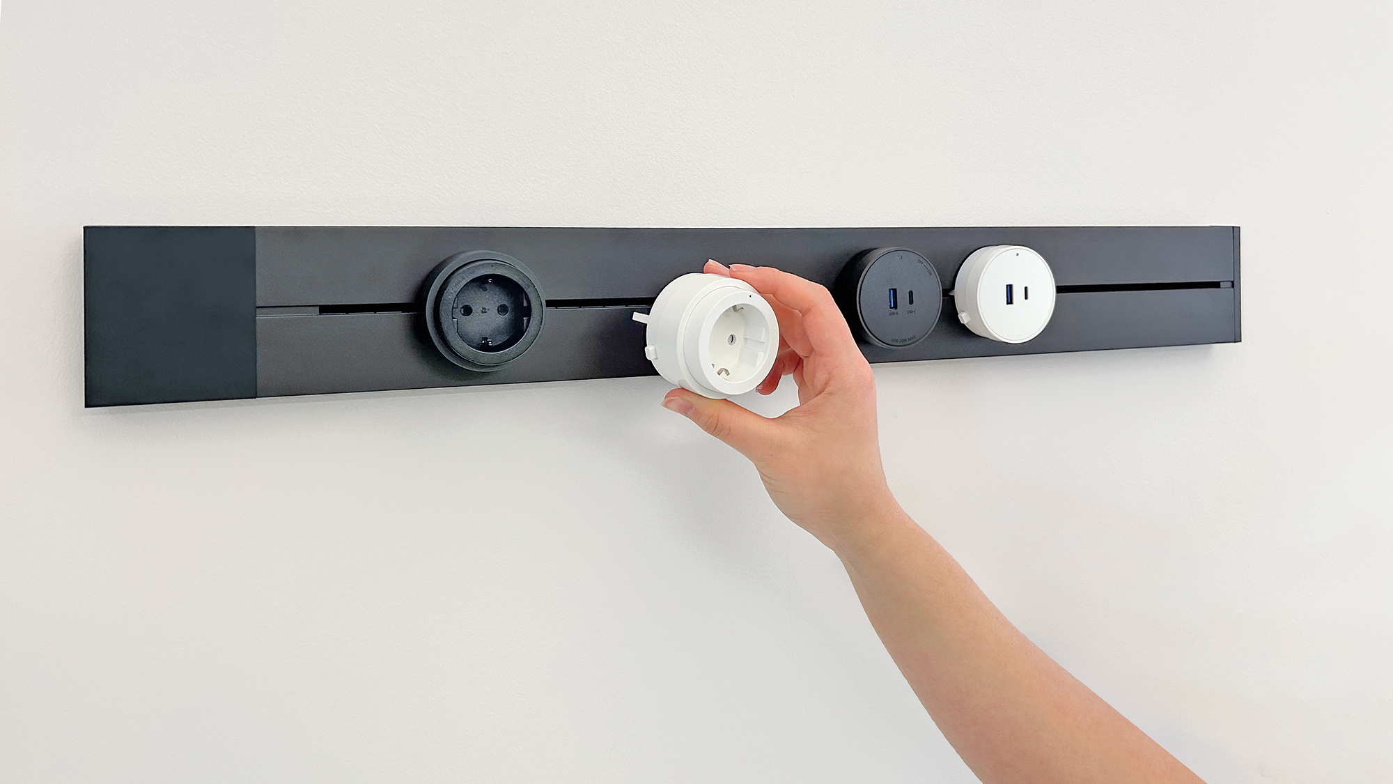 Eine Hand hält eine weiße Steckdose neben schwarzen Steckdosen und einem USB-Anschluss. Alles ist an einem schwarzen Streifen an einer weißen Wand befestigt.