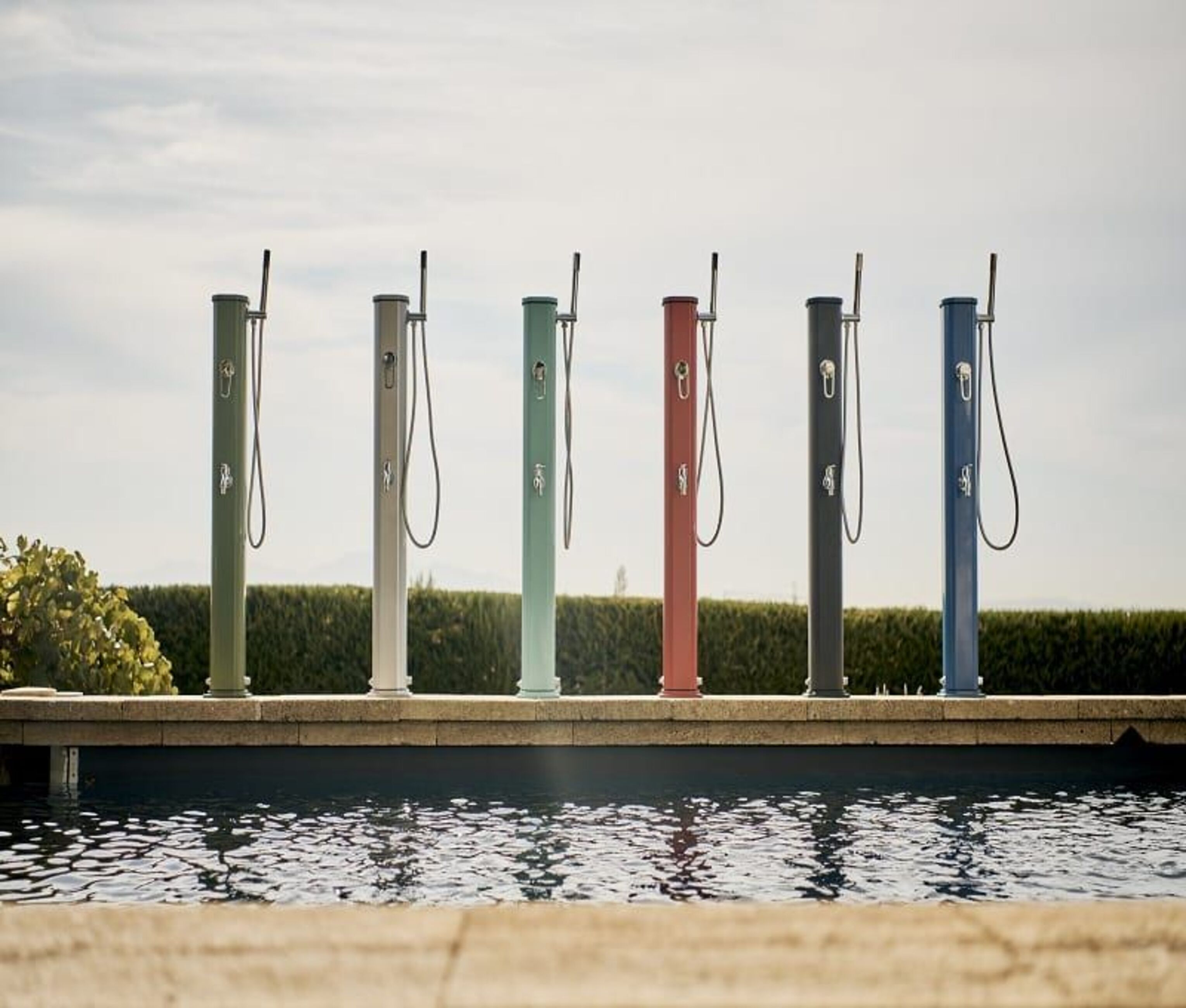 Seis duchas exteriores coloridas junto a una piscina. Verde, gris, turquesa, rojo, gris oscuro y azul. Cielo y vegetación al fondo.