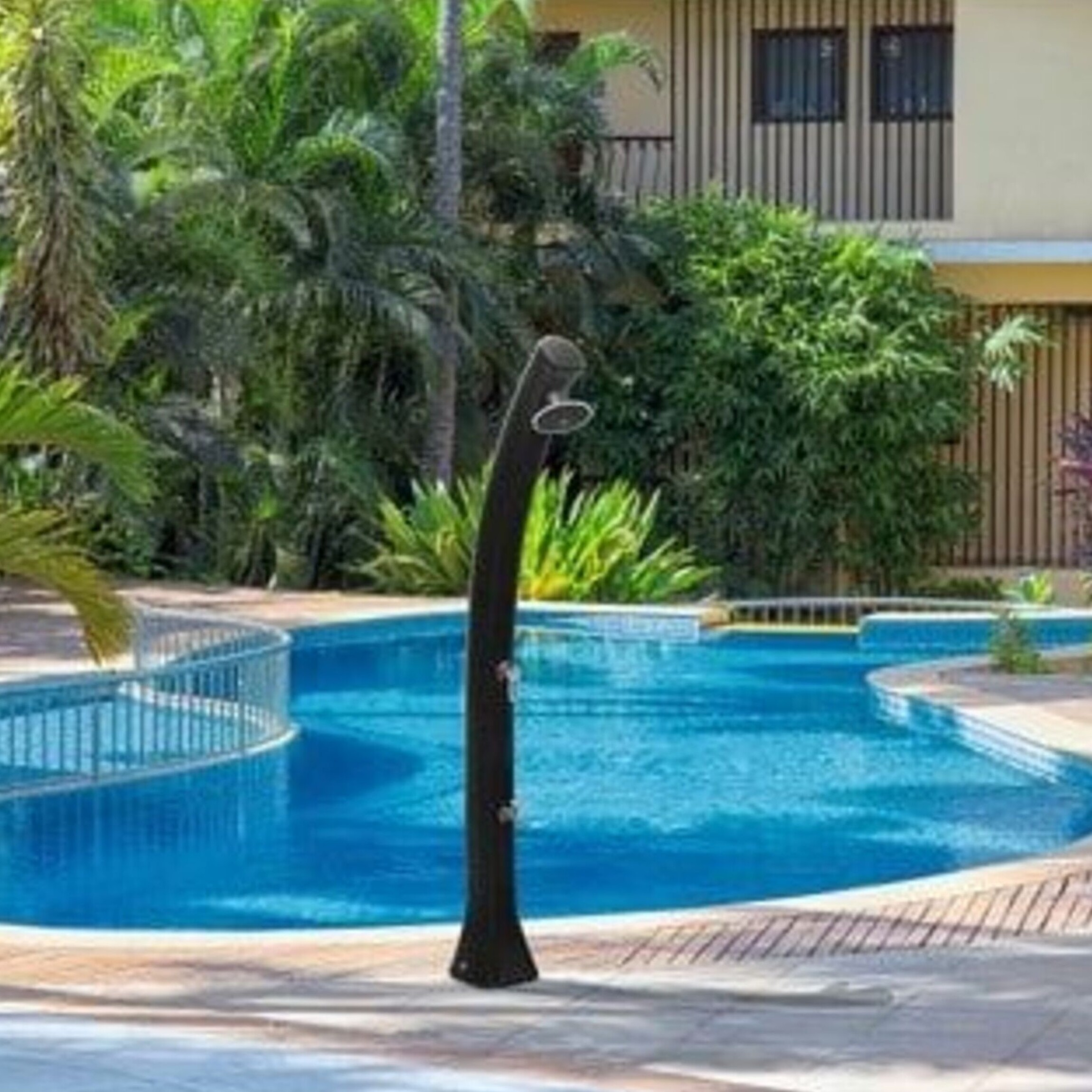 Una escena de piscina al aire libre con una ducha negra. Árboles verdes exuberantes rodean un edificio claro con ventanas. La piscina tiene agua azul.
