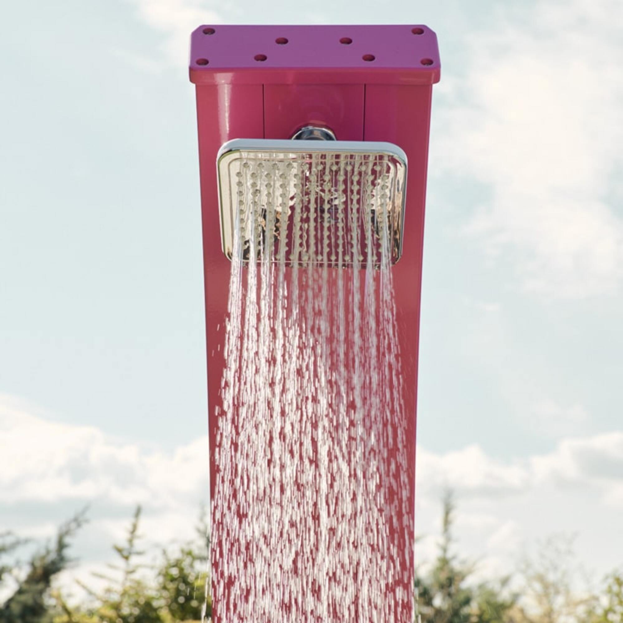 Una ducha al aire libre rosa con agua cayendo. El cielo es azul con nubes y árboles al fondo.