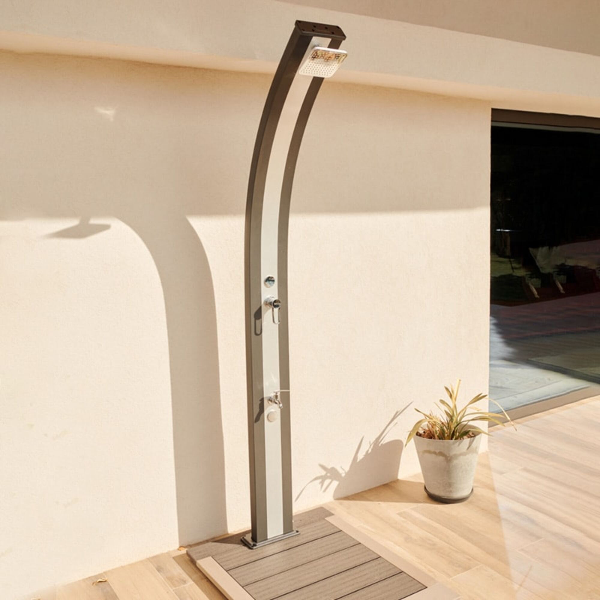 Ducha exterior, gris y blanca, sobre una terraza de madera, junto a una pared crema. Planta en maceta en la terraza. Gran puerta de vidrio al fondo.
