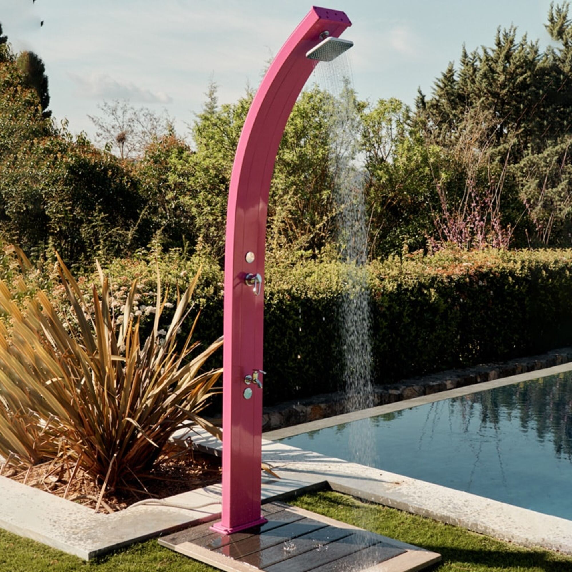 Una ducha rosa al aire libre con agua cayendo. Está en un jardín con piscina y árboles.