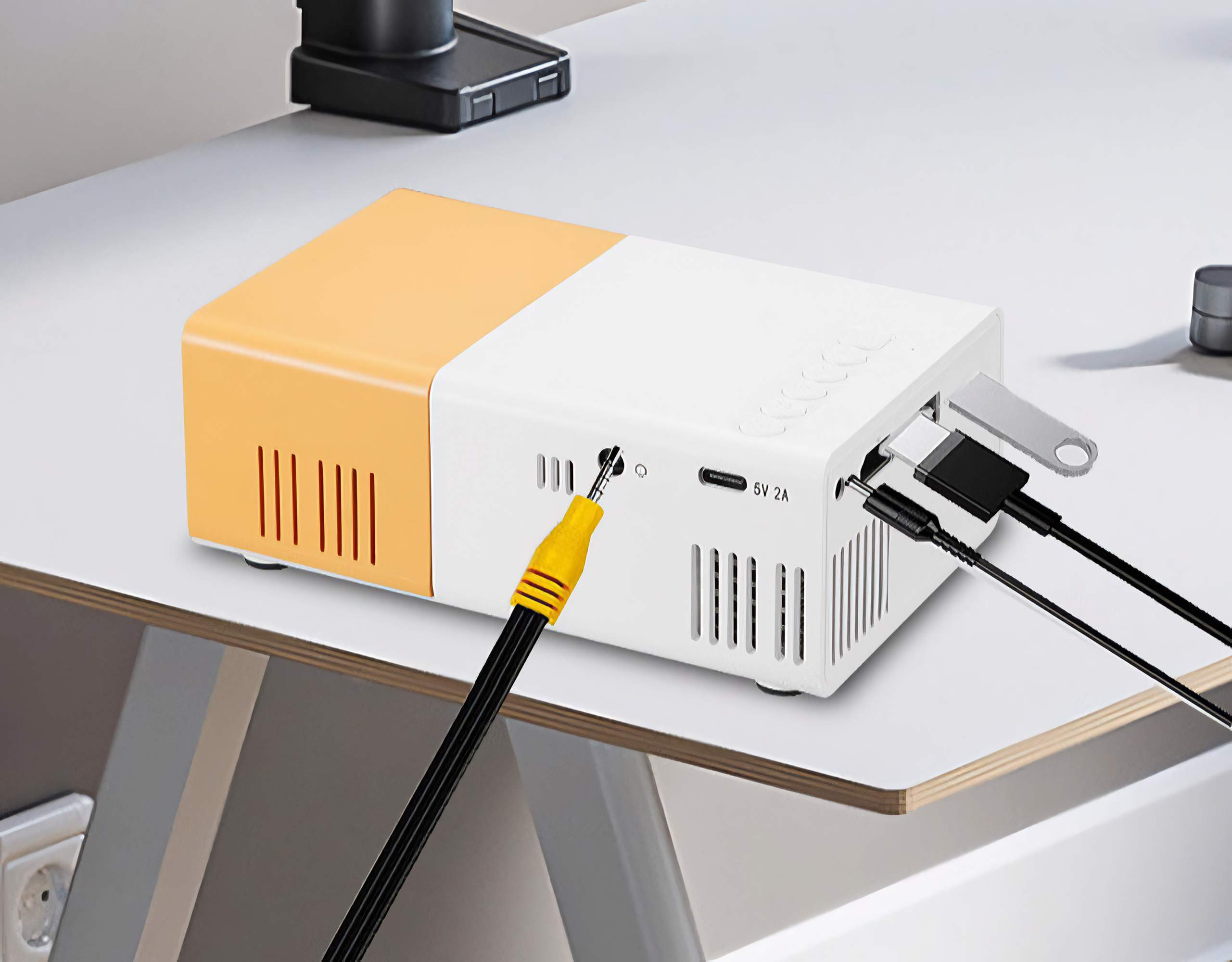 Ein weißer und orangefarbener Projektor mit eingesteckten Kabeln. Ein gelbes Kabel befindet sich vorne. Der Projektor steht auf einer weißen Oberfläche.