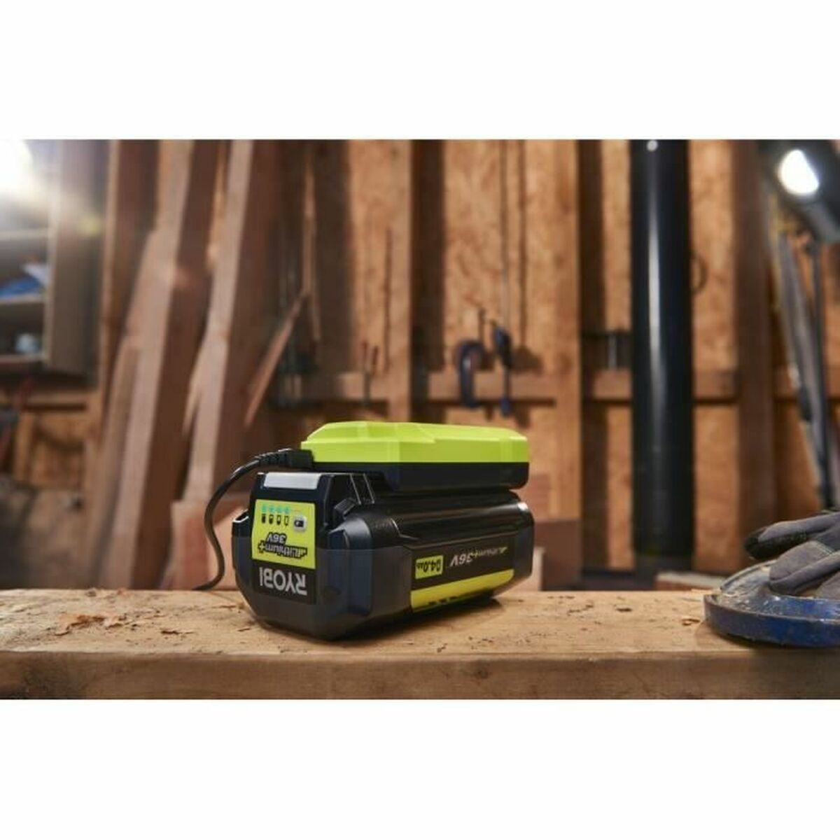 Un paquete de baterías Ryobi negro y amarillo está sobre una superficie de madera. Está enchufado. El fondo es un taller con madera.
