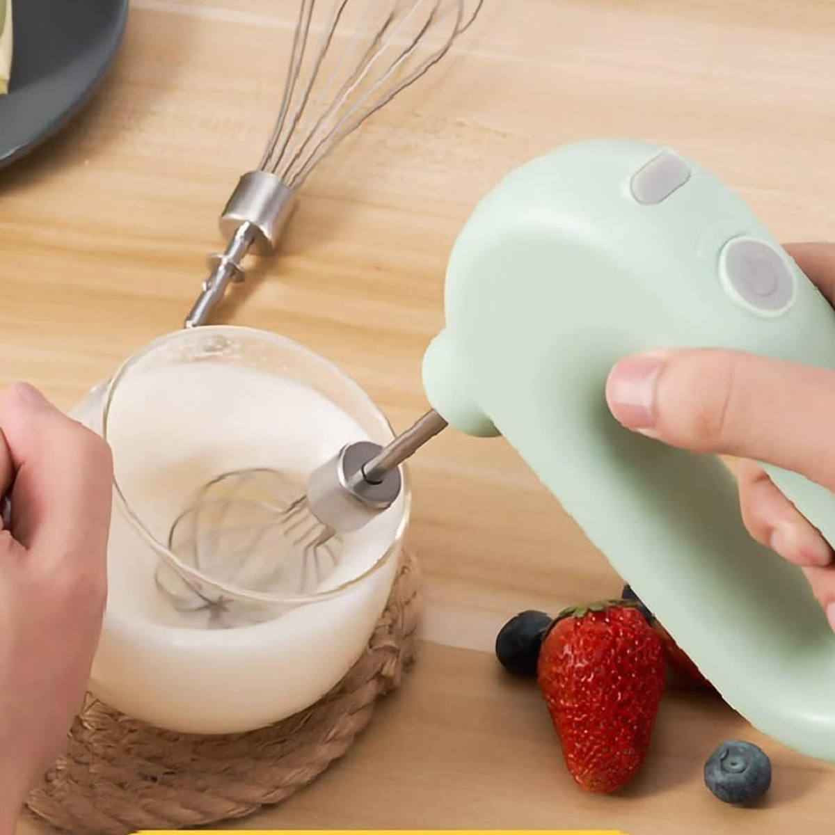 Ein Handmixer verrührt Sahne in einer Glasschüssel. Auf dem Tisch liegen eine Erdbeere und Blaubeeren. Neben der Schüssel steht ein Schneebesen.
