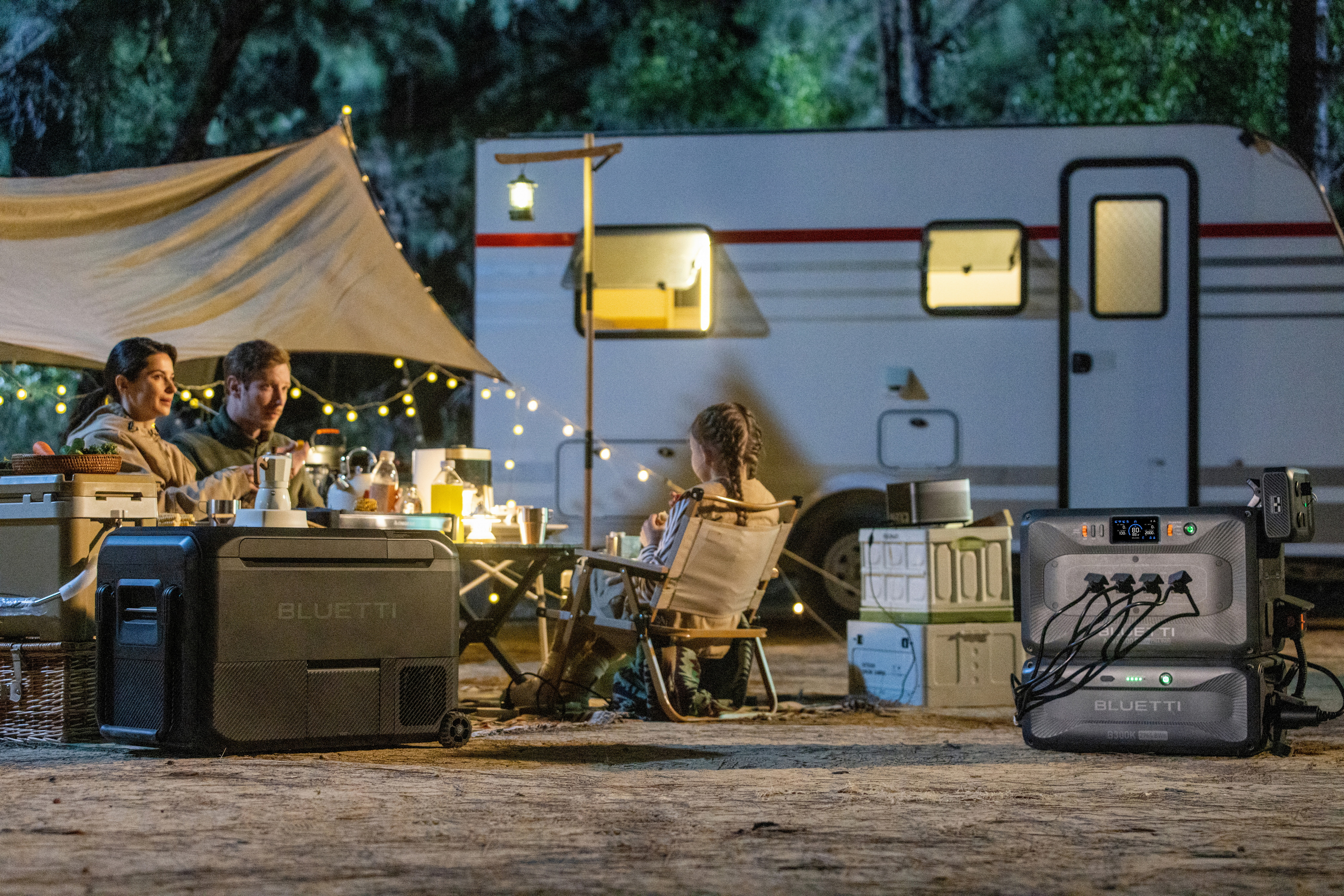 Menschen campen in der Nähe eines Anhängers. Ein Tisch mit Essen und Getränken ist aufgebaut, mit einer Stromstation im Vordergrund.