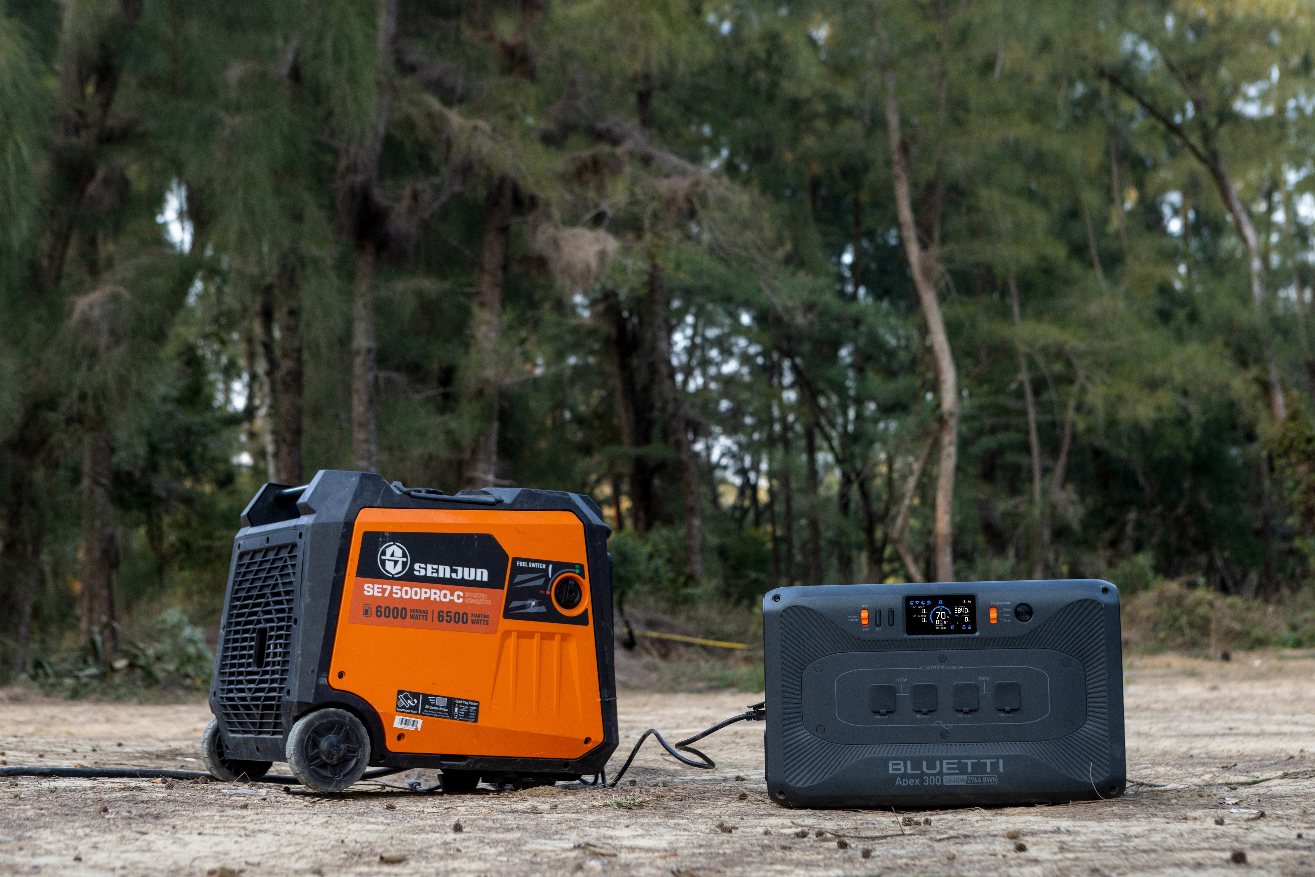 Pomarańczowy i czarny generator oraz szara elektrownia na piaszczystej powierzchni, drzewa w tle.