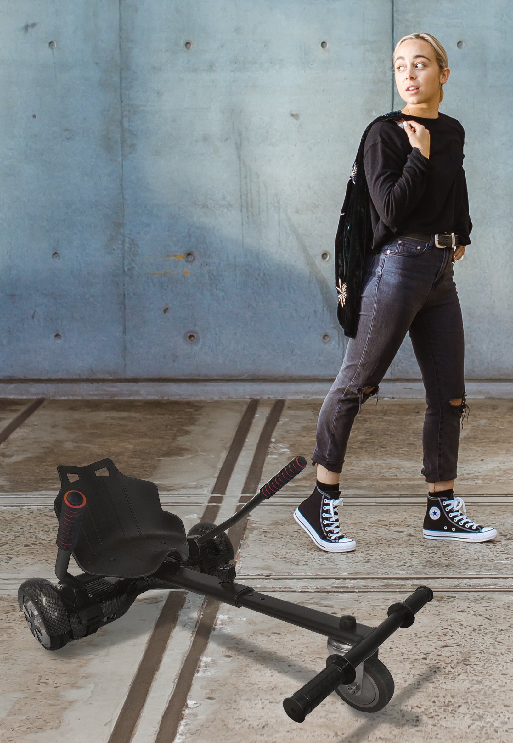 Eine Frau in schwarzen Jeans und Oberteil geht an einem schwarzen, tief liegenden Roller vorbei. Betonwand im Hintergrund.
