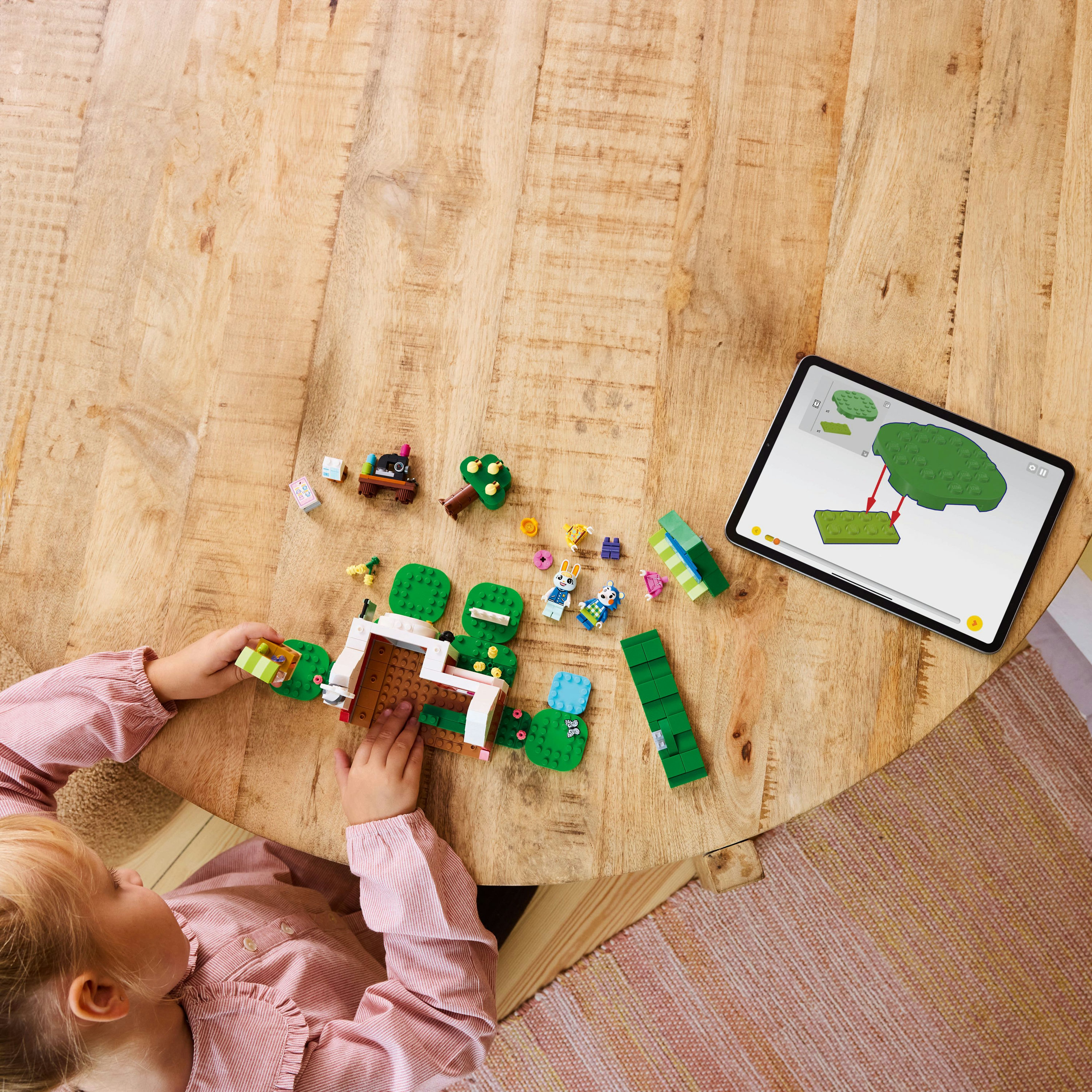 Dziecko składa klocki Lego na drewnianym stole, kierując się instrukcjami z tabletu. Różne elementy Lego są rozrzucone.