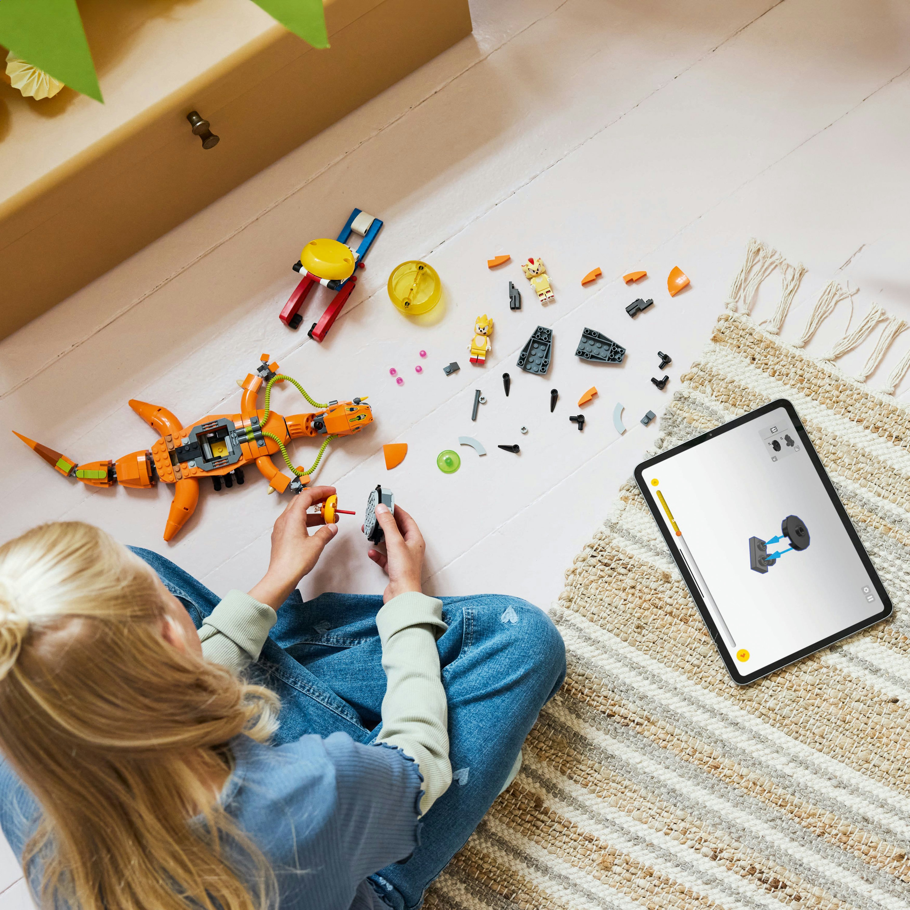 Osoba składa Lego. Pomarańczowa Lego istota i klocki na podłodze. Tablet wyświetla instrukcje budowy.