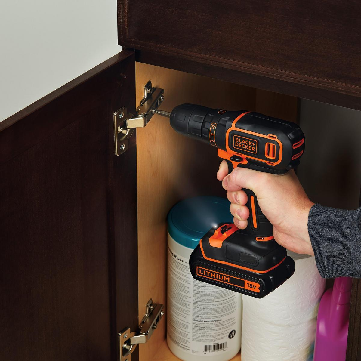 Osoba używa wiertarki Black+Decker do zamontowania zawiasu na brązowych drzwiach szafki. W szafce znajdują się środki czystości.