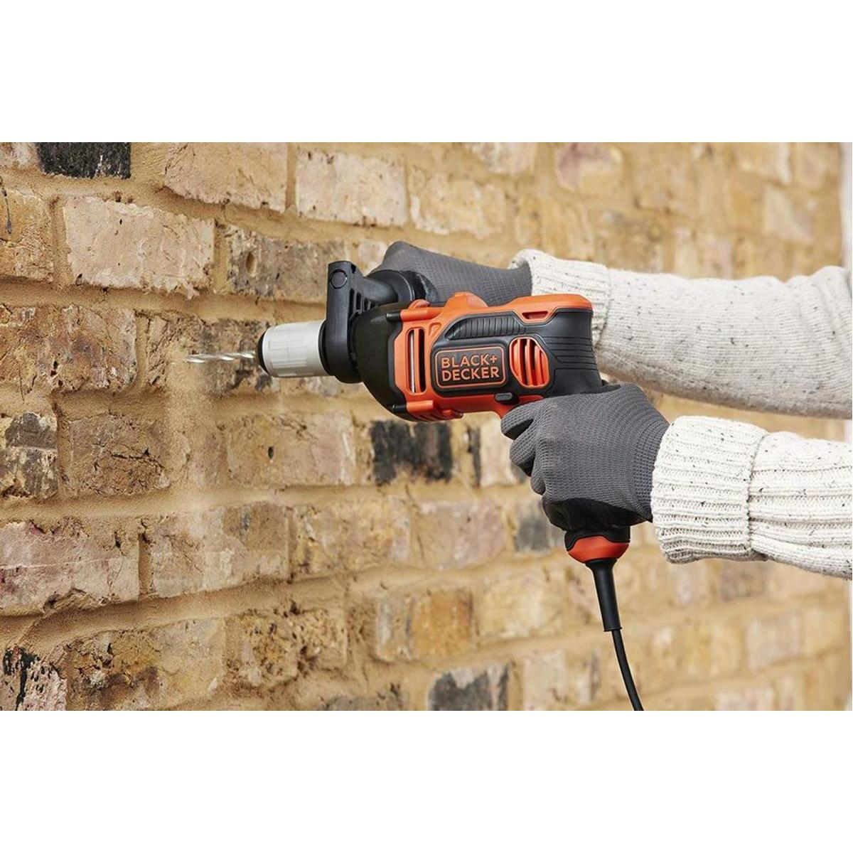 Una persona taladra una pared de ladrillos con una herramienta Black+Decker. La herramienta es naranja y negra. La persona lleva guantes y un suéter.