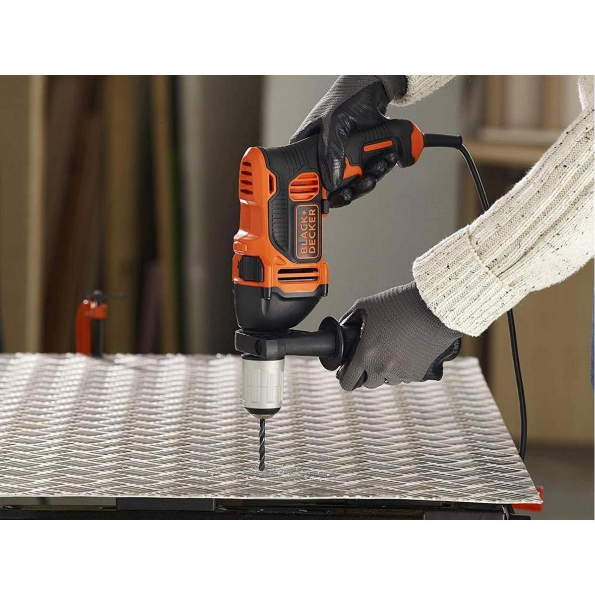 Una persona taladra una lámina de metal con un taladro Black+Decker. El taladro es naranja y negro. La persona usa guantes y un suéter.
